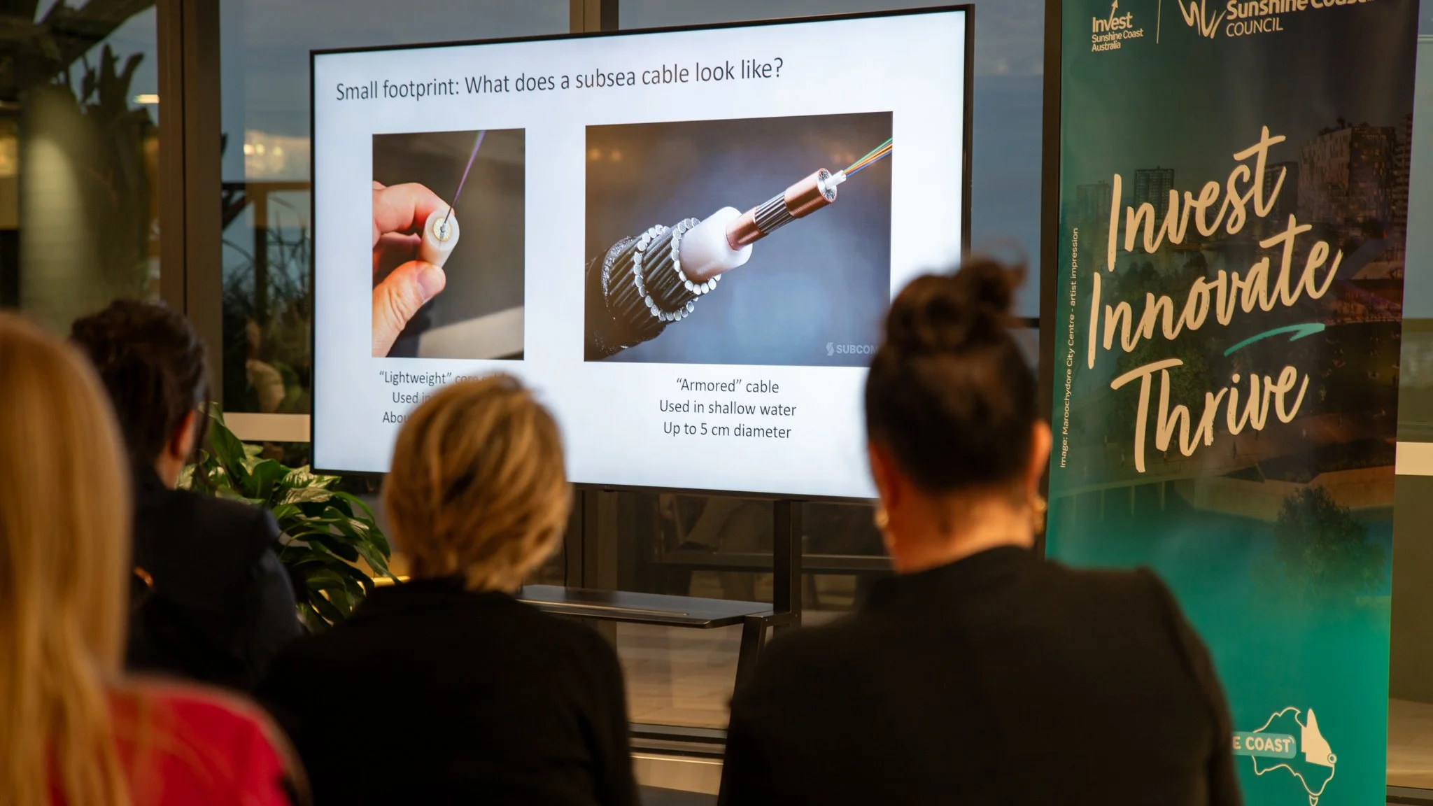 People attending a presentation in front of a screen showing images and information about subsea cables, including a close-up of two different types of cables with descriptive labels.