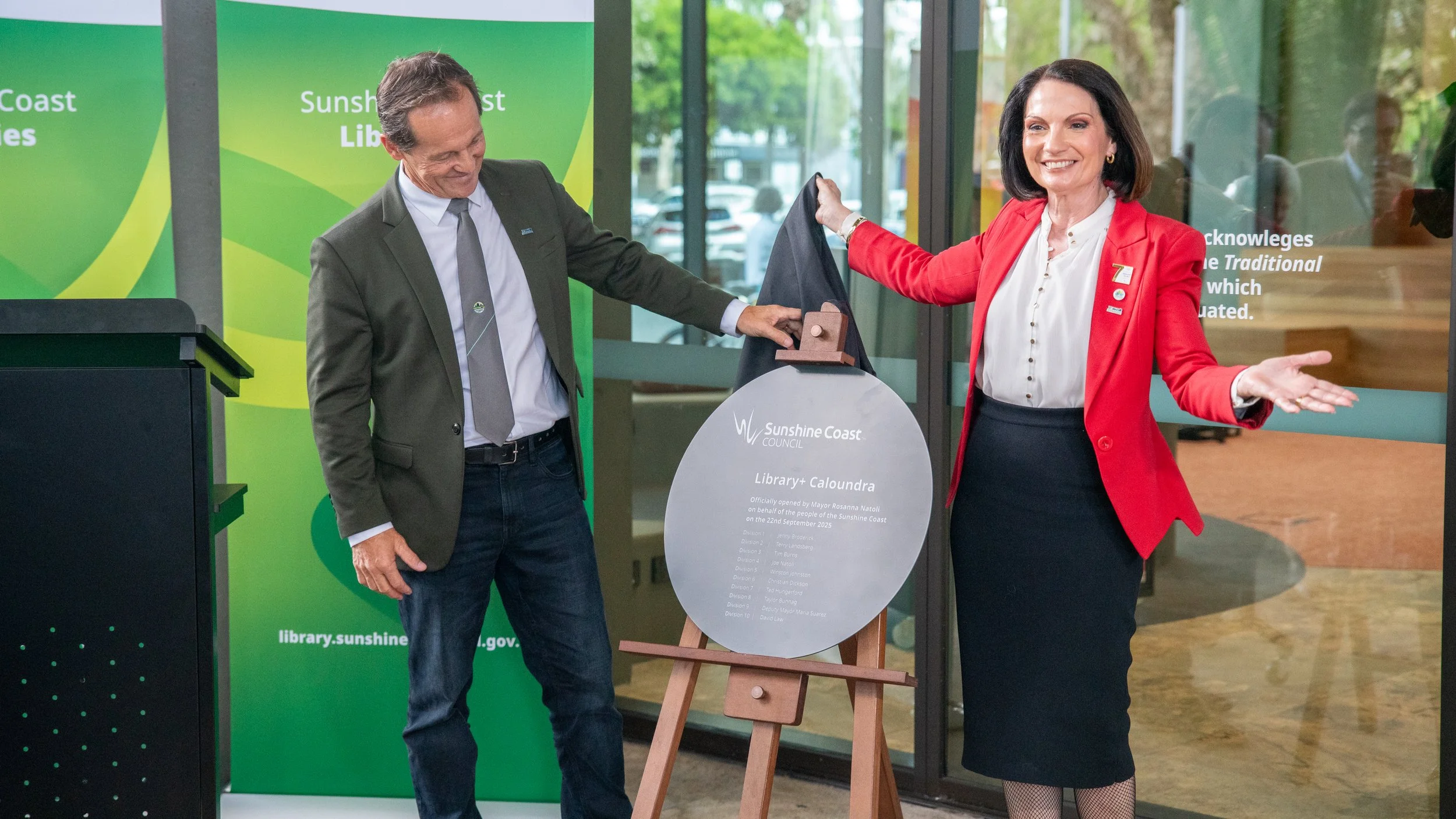 Two people unveil a plaque at a Sunshine Coast Library opening. The man wears a dark jacket and jeans, and the woman wears a red blazer and a dark skirt. They are smiling and standing in front of green and glass backgrounds.