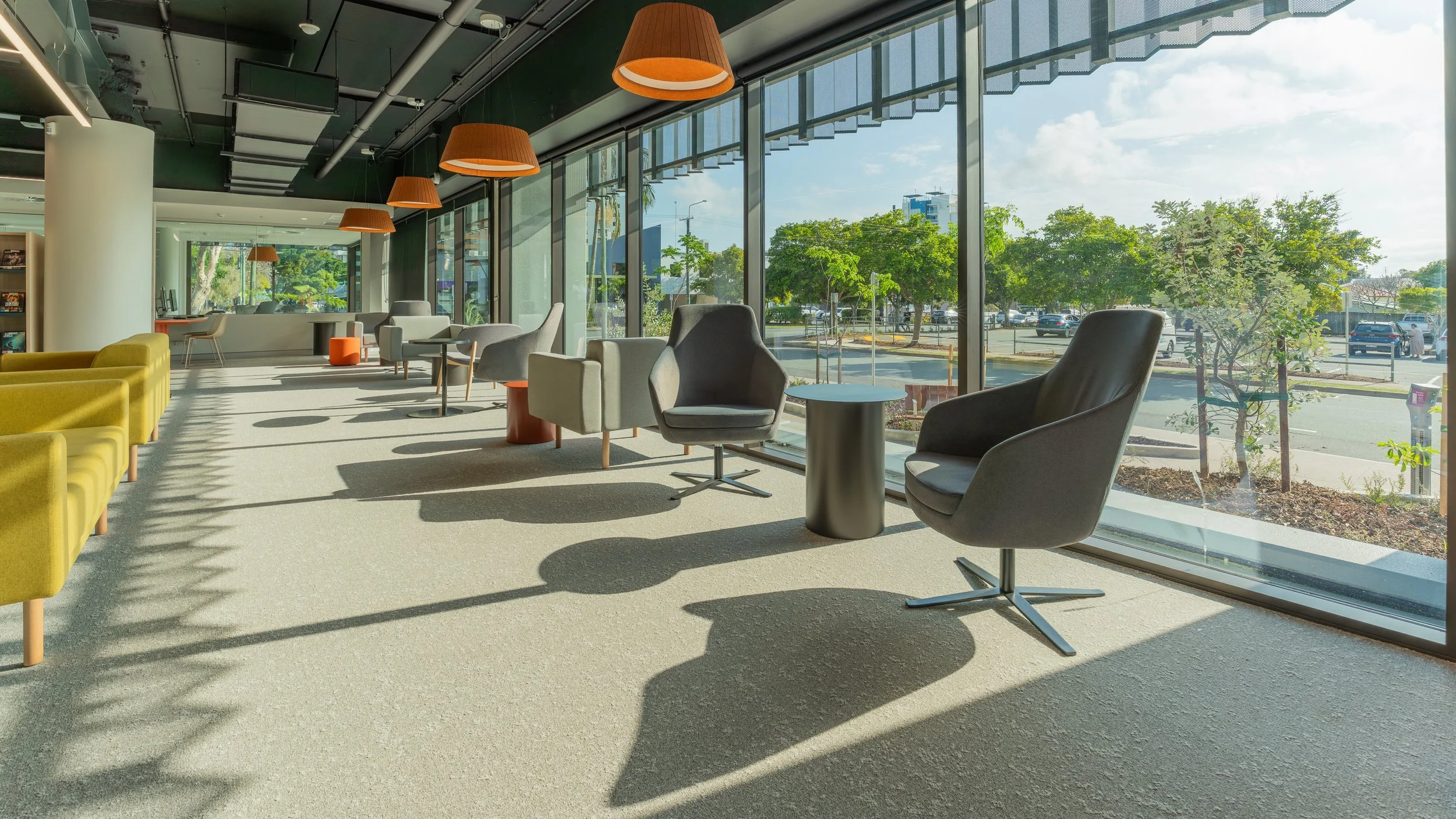 Bright modern lounge area with large windows, overlooking a parking lot and green trees, furnished with various armchairs, side tables, and hanging orange lamps.