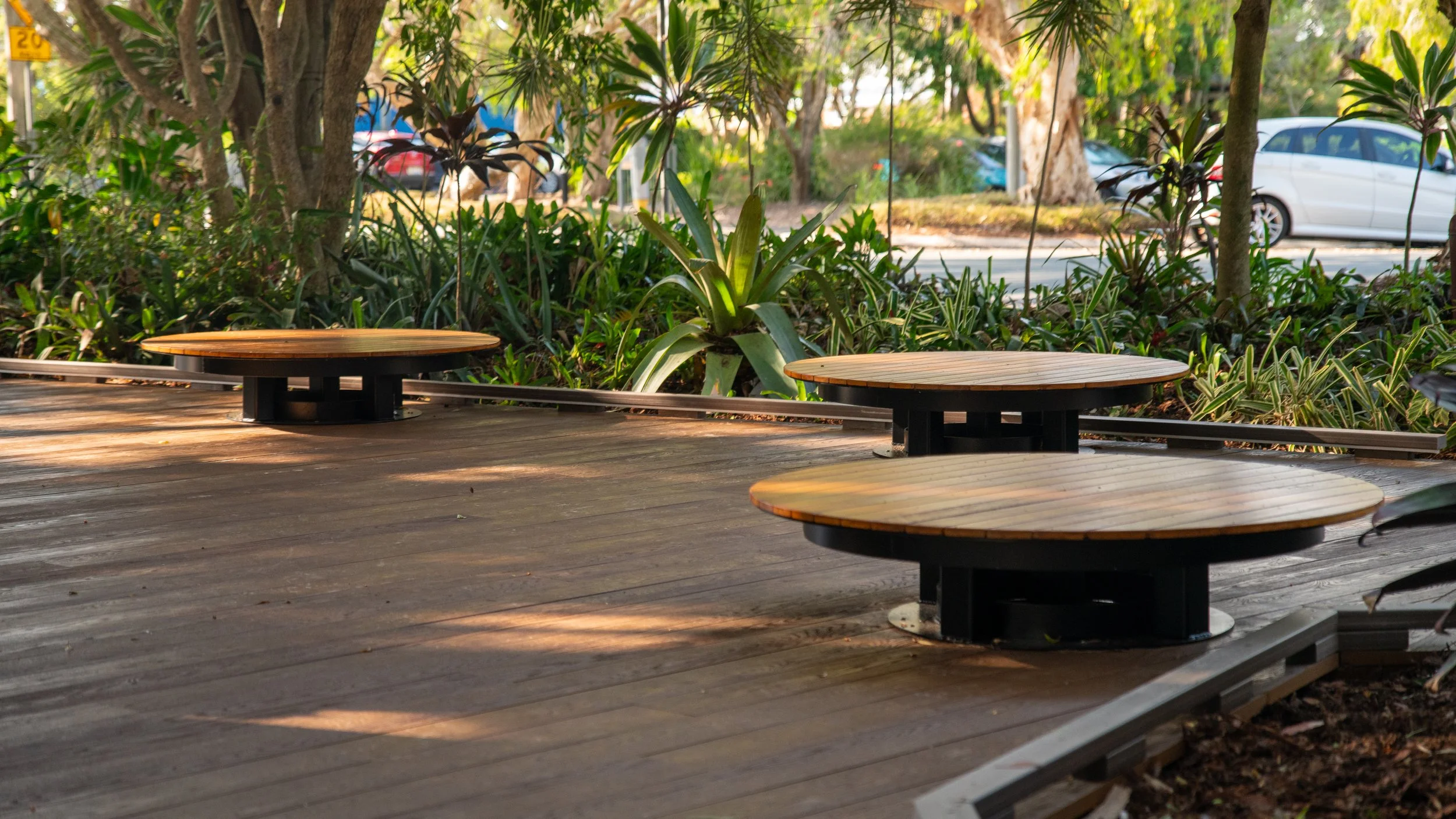 Three round wooden platform seats on a wooden deck in a lush, green outdoor setting with plants and trees, and parked cars in the background.