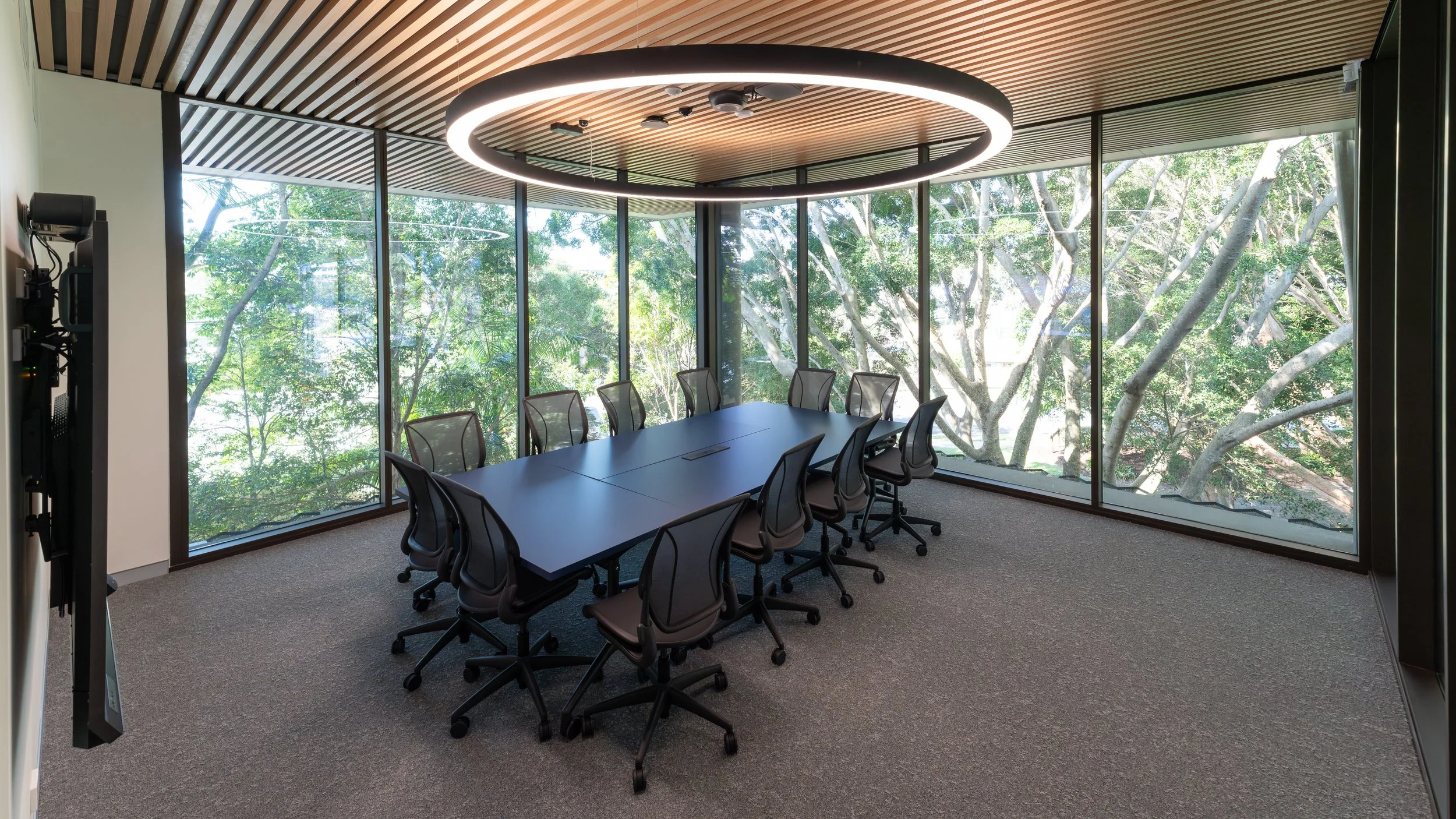 Modern conference room with a long table, 10 chairs, large windows showing trees outside, and a circular ceiling light.