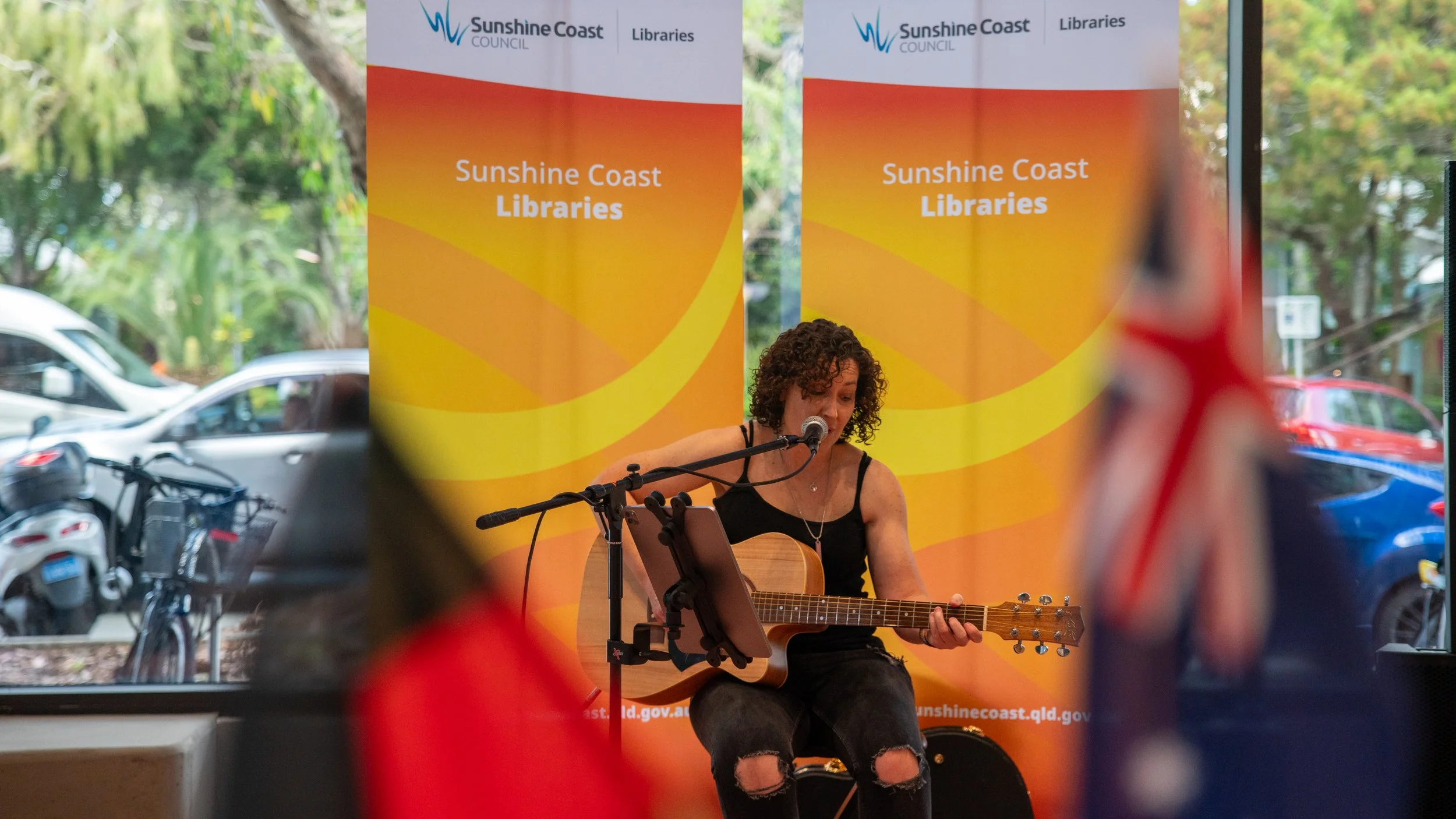 A woman with curly hair wearing a black tank top and ripped jeans is playing an acoustic guitar and singing into a microphone at a Sunshine Coast Libraries event, with banners and parked cars visible in the background.