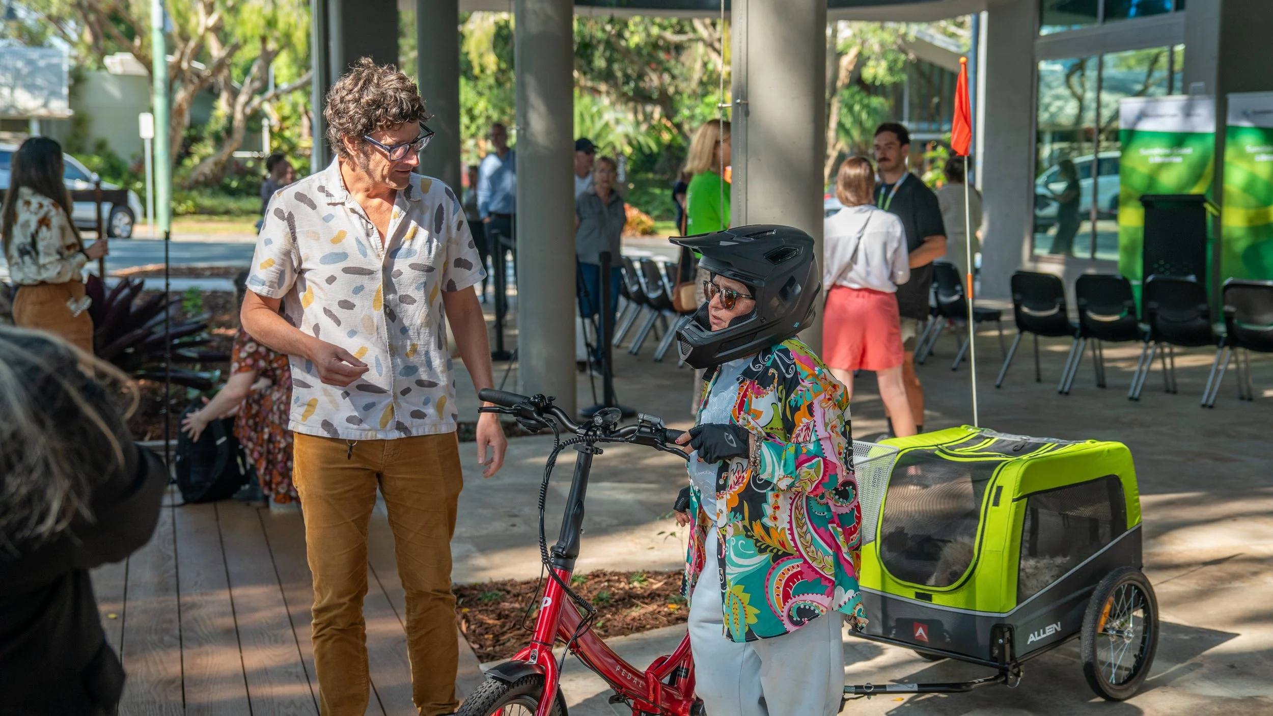 A woman with sunglasses and a colorful jacket stands next to a pedal bicycle with a trailer attached, talking to a man with curly hair, glasses, and a patterned shirt, in an outdoor setting with several people and chairs in the background.