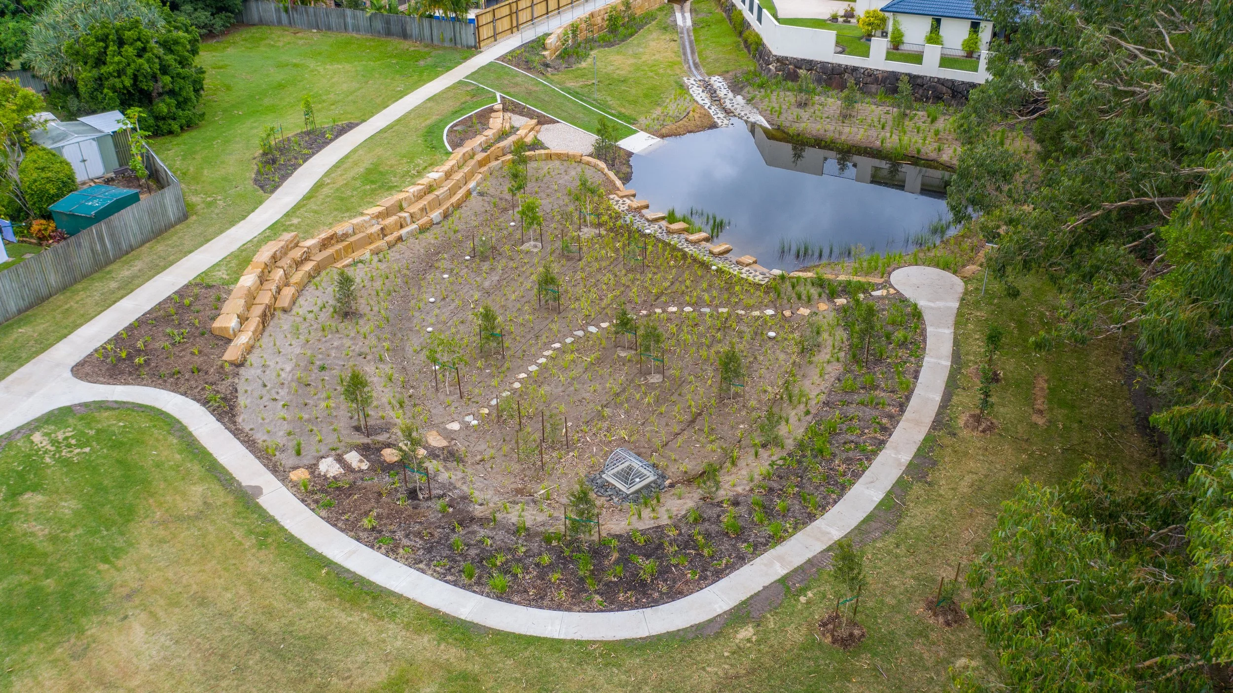 Aerial view of a landscaped backyard featuring a pond, winding concrete pathways, a garden with young plants and trees, and a stone retaining wall. There are neighboring houses and lush greenery surrounding the area.