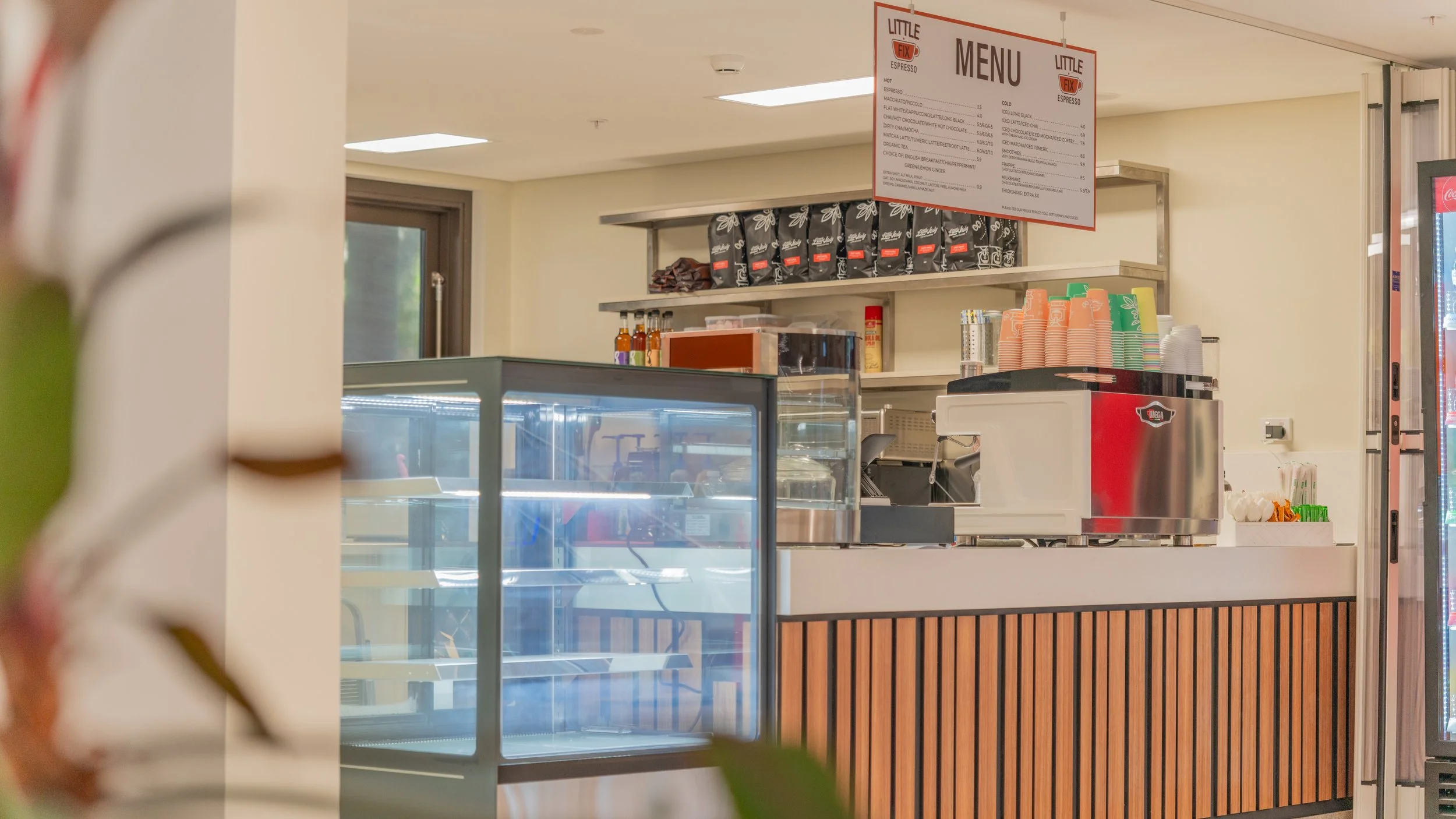Empty display case and a coffee counter with cups, a coffee machine, and a "Menu" sign inside a cafe or coffee stand.