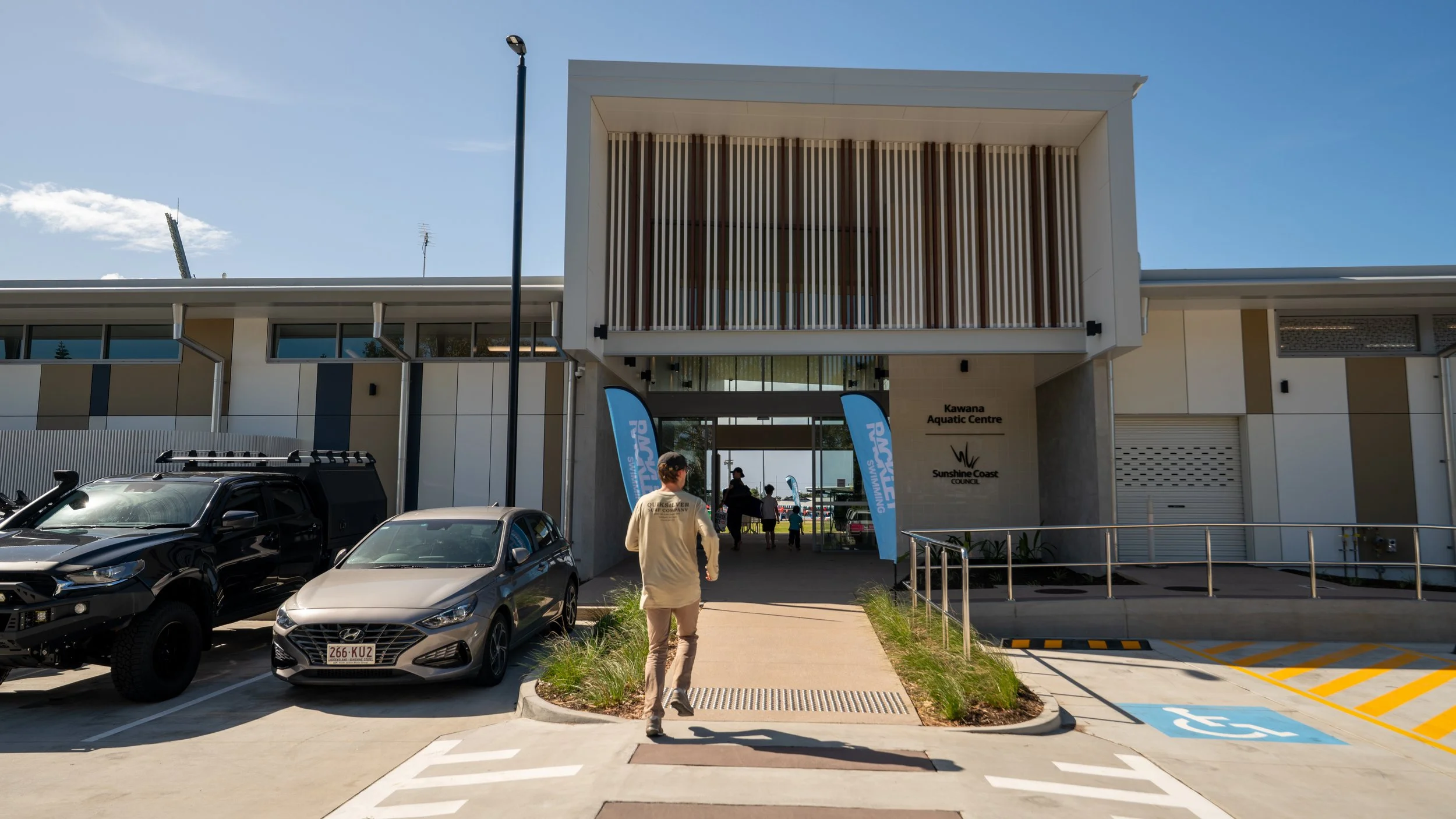 Front entrance of Kawana Aquatic Centre with vehicles parked outside and flags that read 'PMAR Swimming'.