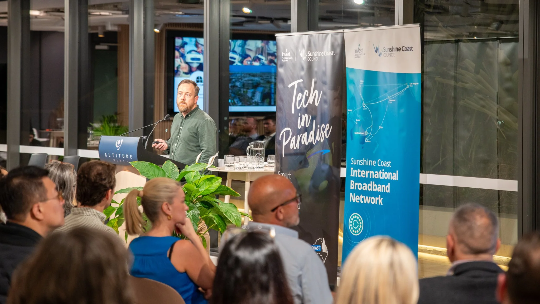 A man giving a presentation at a conference with a screen behind him, titled 'Tech in Paradise', with an audience watching, indoors with large windows, plants, and promotional banners for Sunshine Coast.