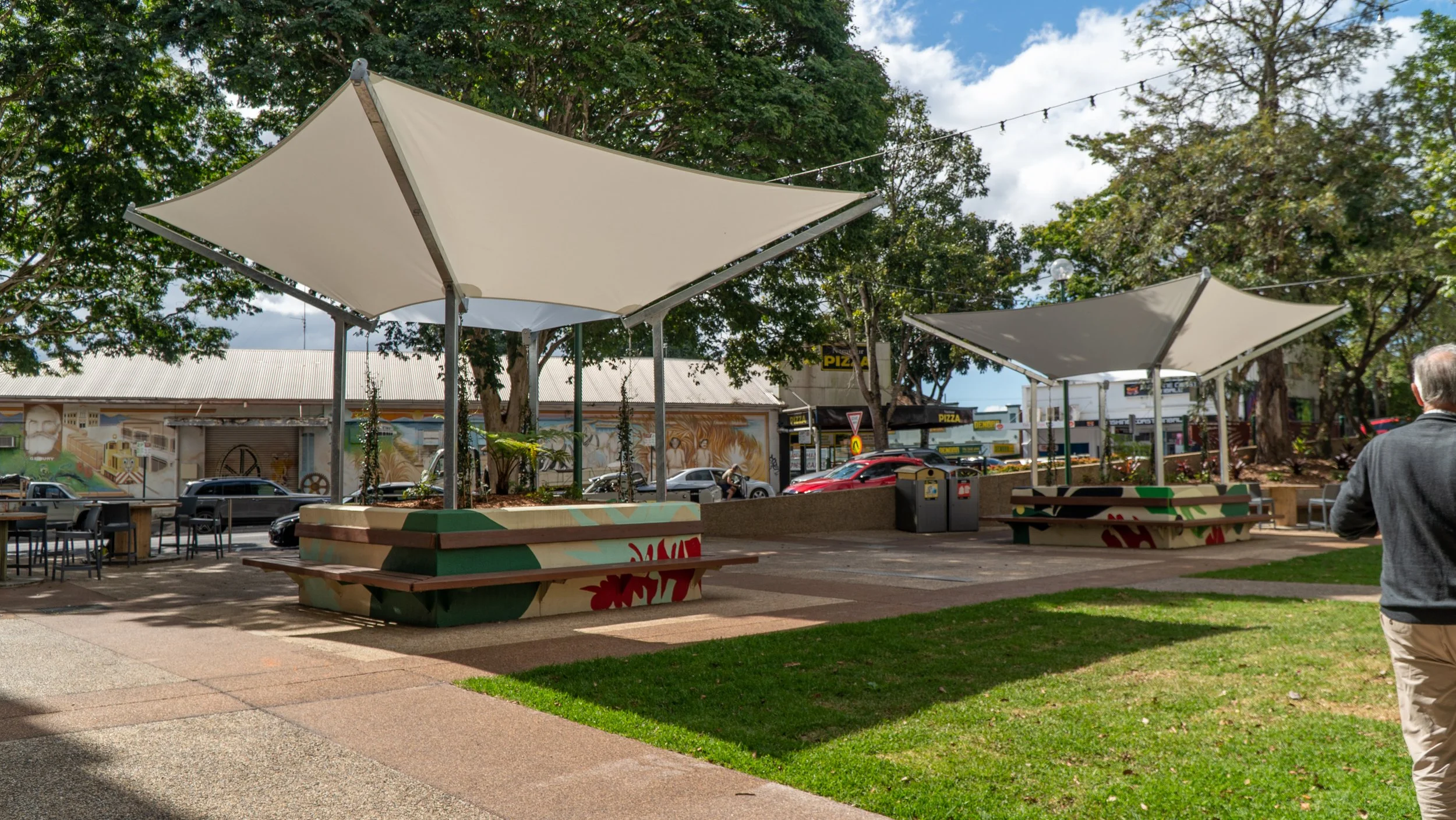 Outdoor seating area with two large white shade structures, some tables and chairs, and a man walking on the sidewalk. In the background, there are trees, a mural painted on a building wall, cars parked, and a pizza restaurant sign.