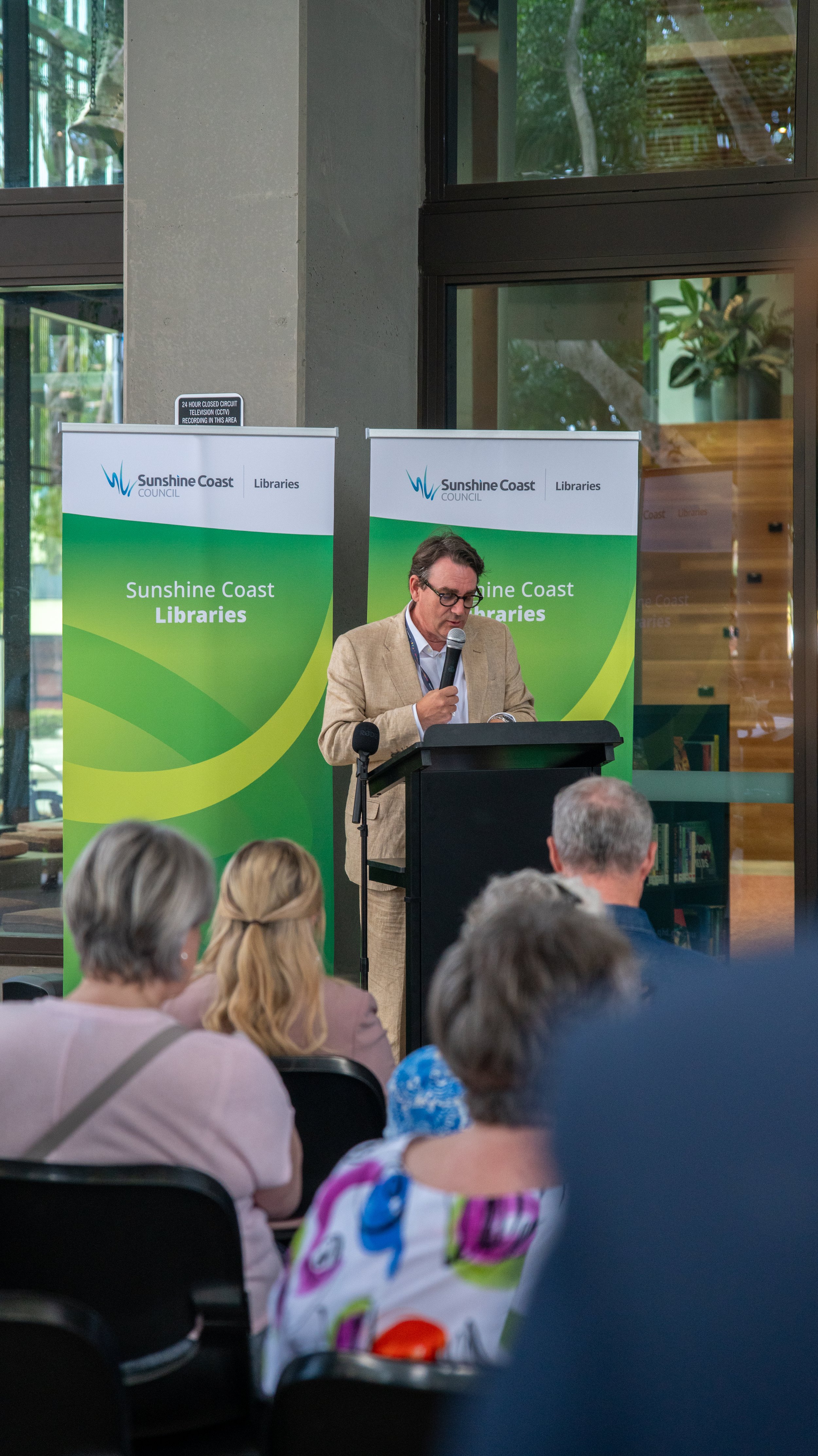 A man in a beige suit and glasses is speaking into a microphone at a podium during a presentation at Sunshine Coast Libraries event, with an audience of diverse adults seated in front, inside a modern building with large windows and green scenery out