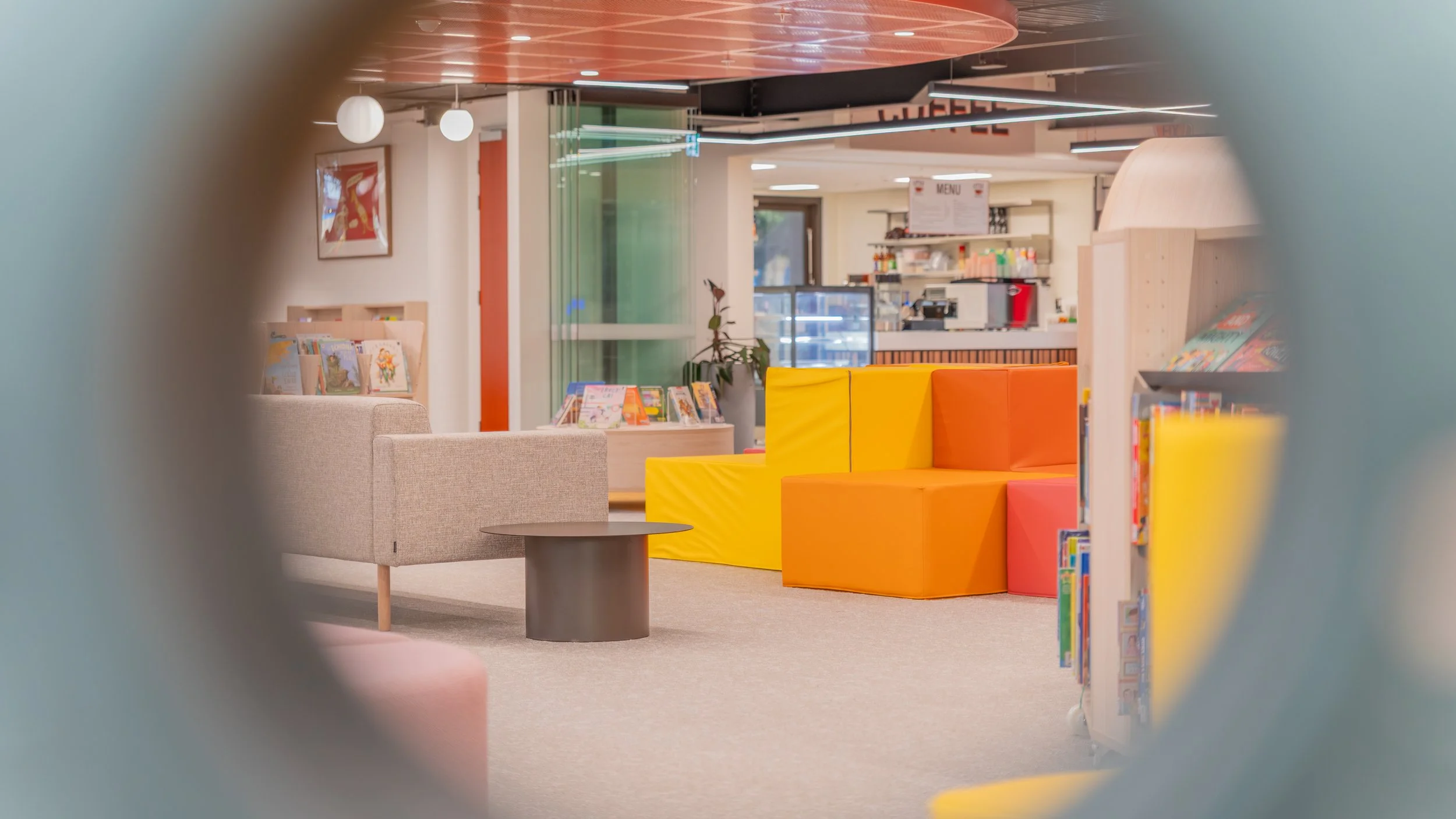 A colorful children's area in a library, featuring yellow and orange padded seating, bookshelves, and a beige armchair, with a view of the cafe and menu in the background.