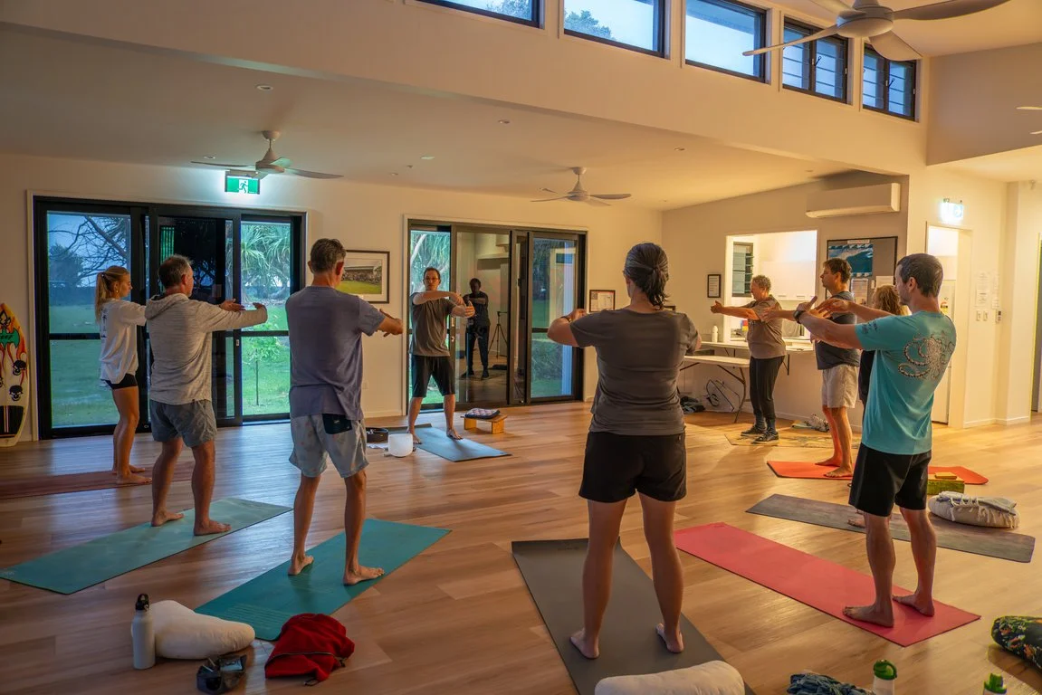 Group of people participating in a yoga class in a spacious, well-lit room with wooden floors and large windows.