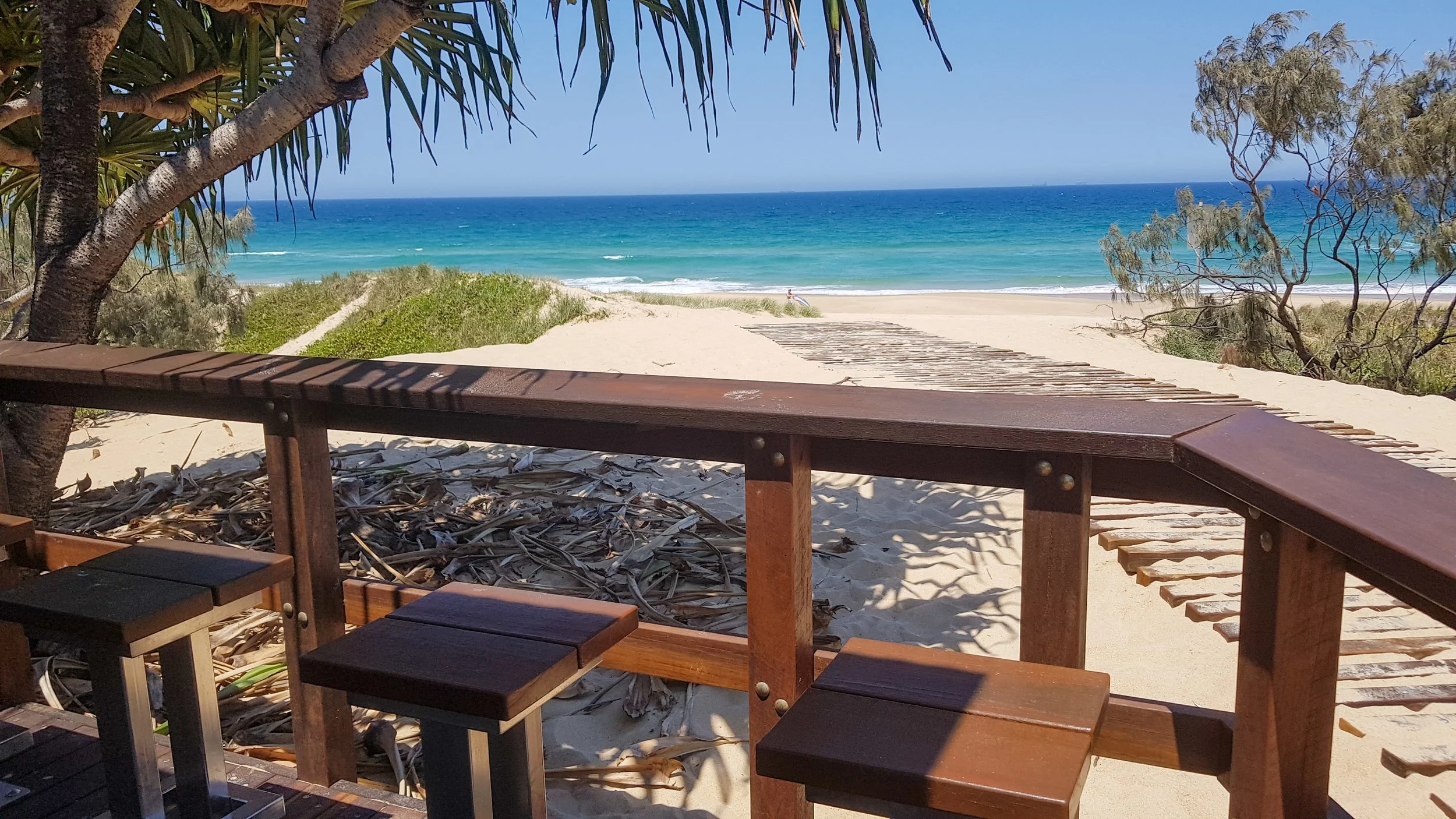 View of a sunny beach from a wooden deck, with trees on either side, a sandy pathway, and the ocean in the background.