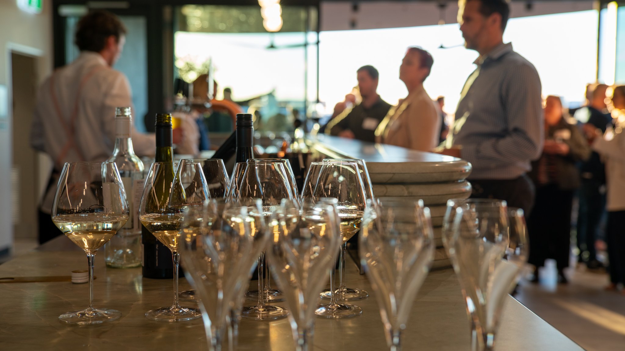 Several wine glasses filled with white wine and bottles on a wooden bar counter, with people socializing in the background inside a bright, modern venue.