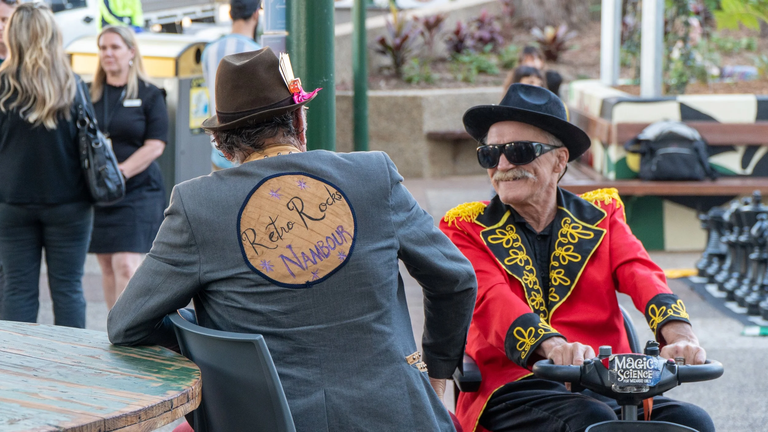 Two older men dressed in retro clothing, one with a brown hat and the other with a black hat and sunglasses, sitting at an outdoor event. The man with the brown hat has a patch on his jacket that reads 'Retro Rocks Nambour.' The man with the black ha