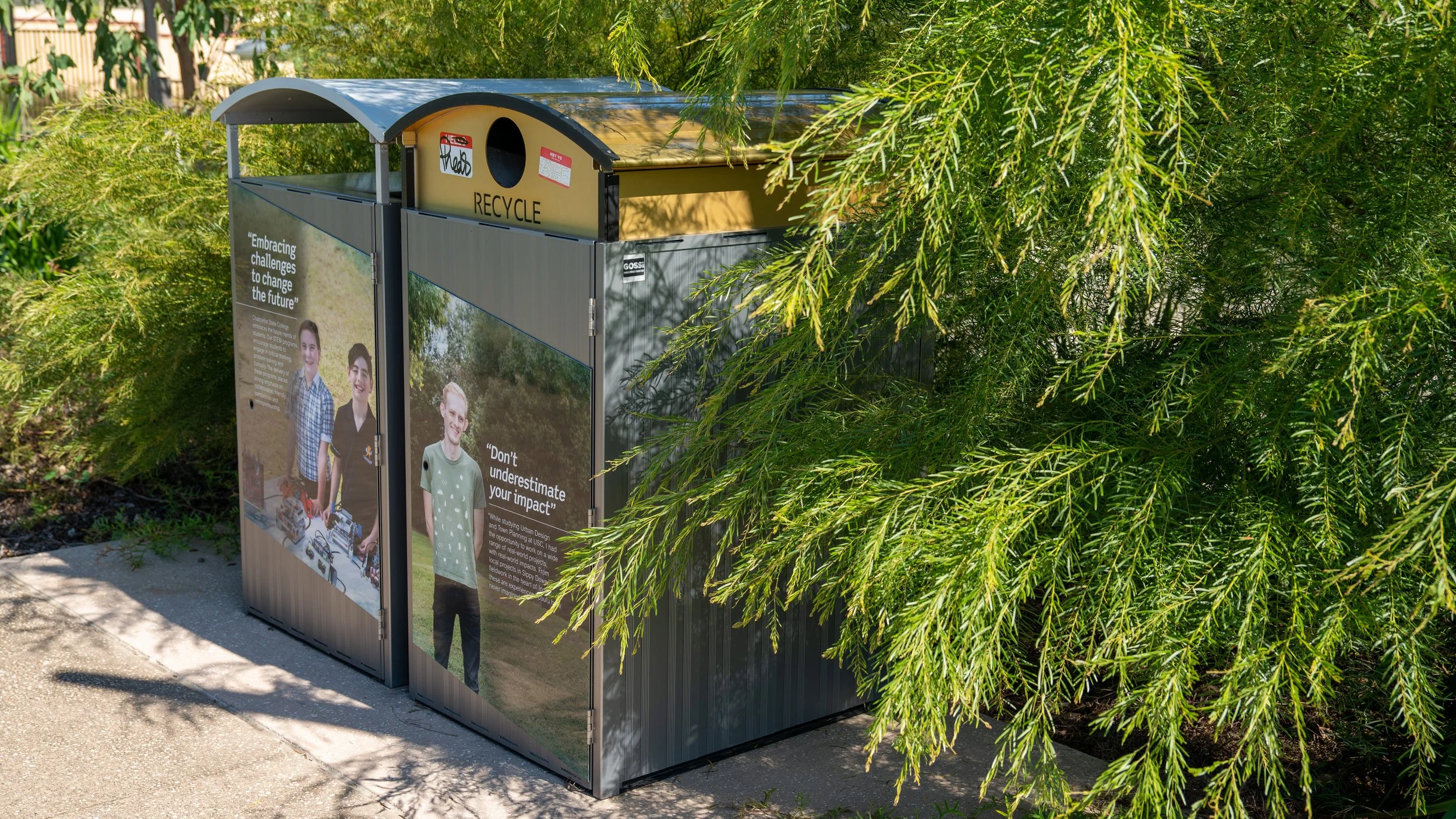 Outdoor recycling bin with a gray metal structure, featuring two panels with photos and quotes, and a yellow lid labeled 'RECYCLE', partially obscured by green foliage.