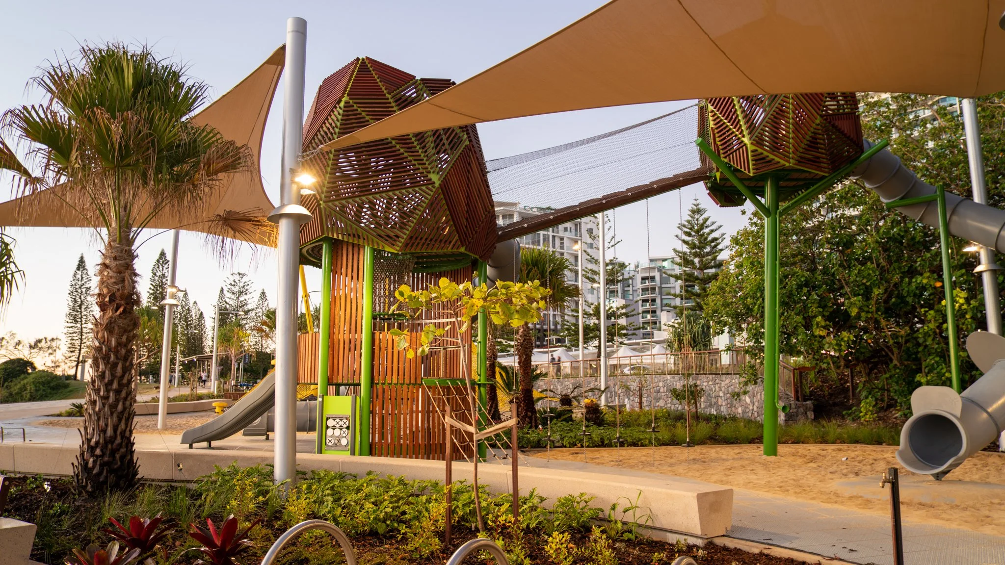 Children's playground with slides, climbing structures, and shade sails, surrounded by trees and high-rise buildings.
