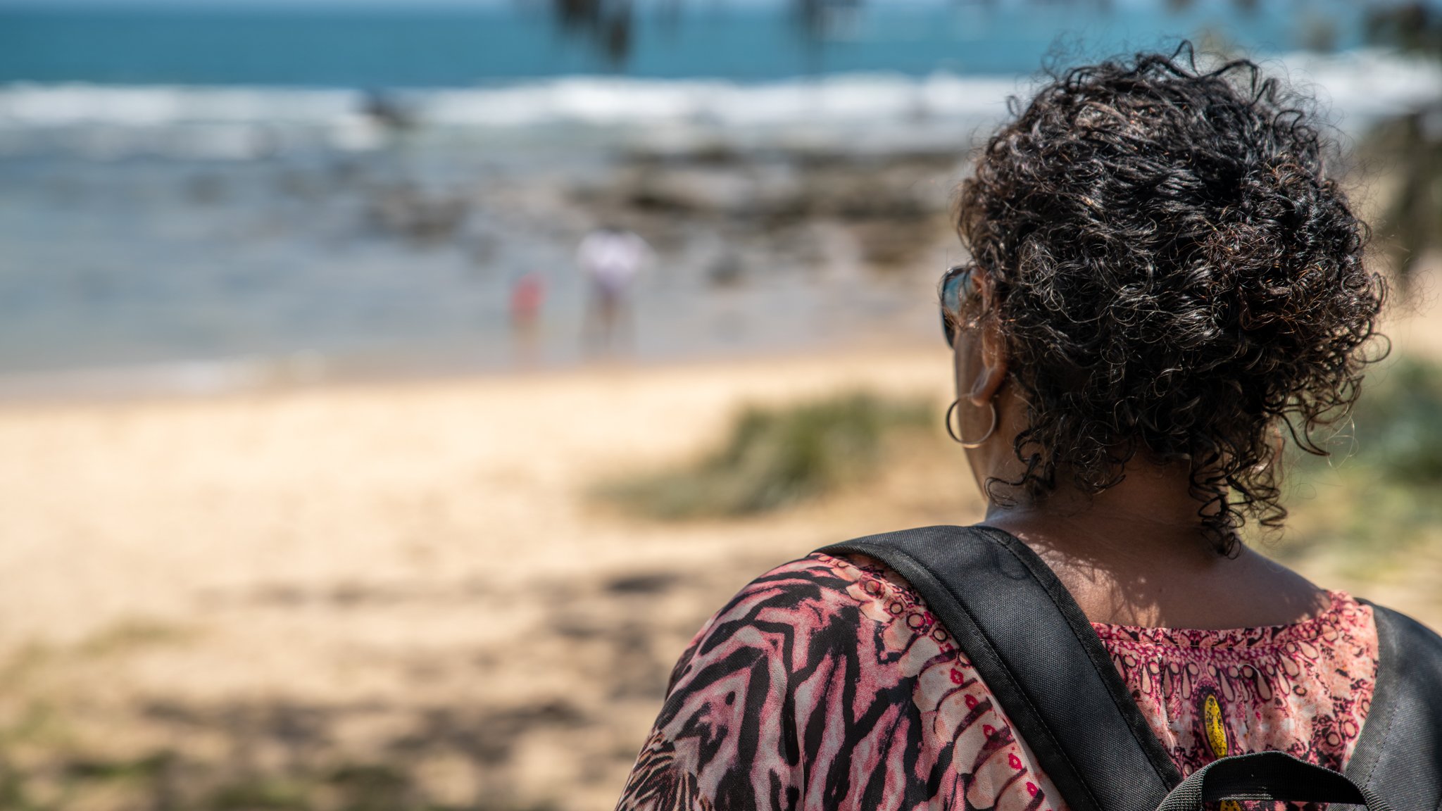 A woman with curly hair and hoop earrings gazes towards the beach and ocean while wearing a patterned top and carrying a backpack.