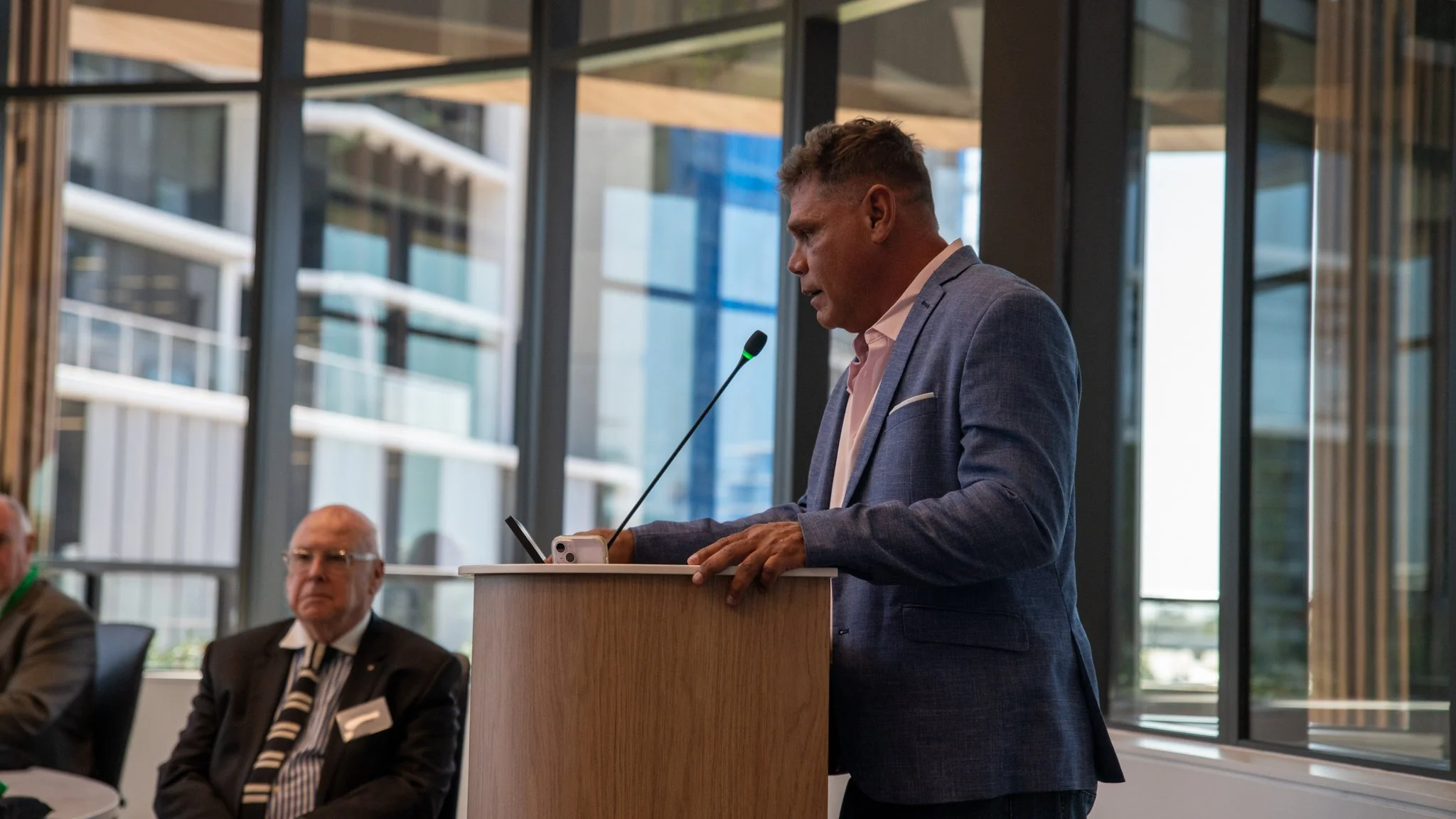 A man in a blue suit speaking at a podium during a meeting, with two other men seated in the background, in a modern office building with large windows.