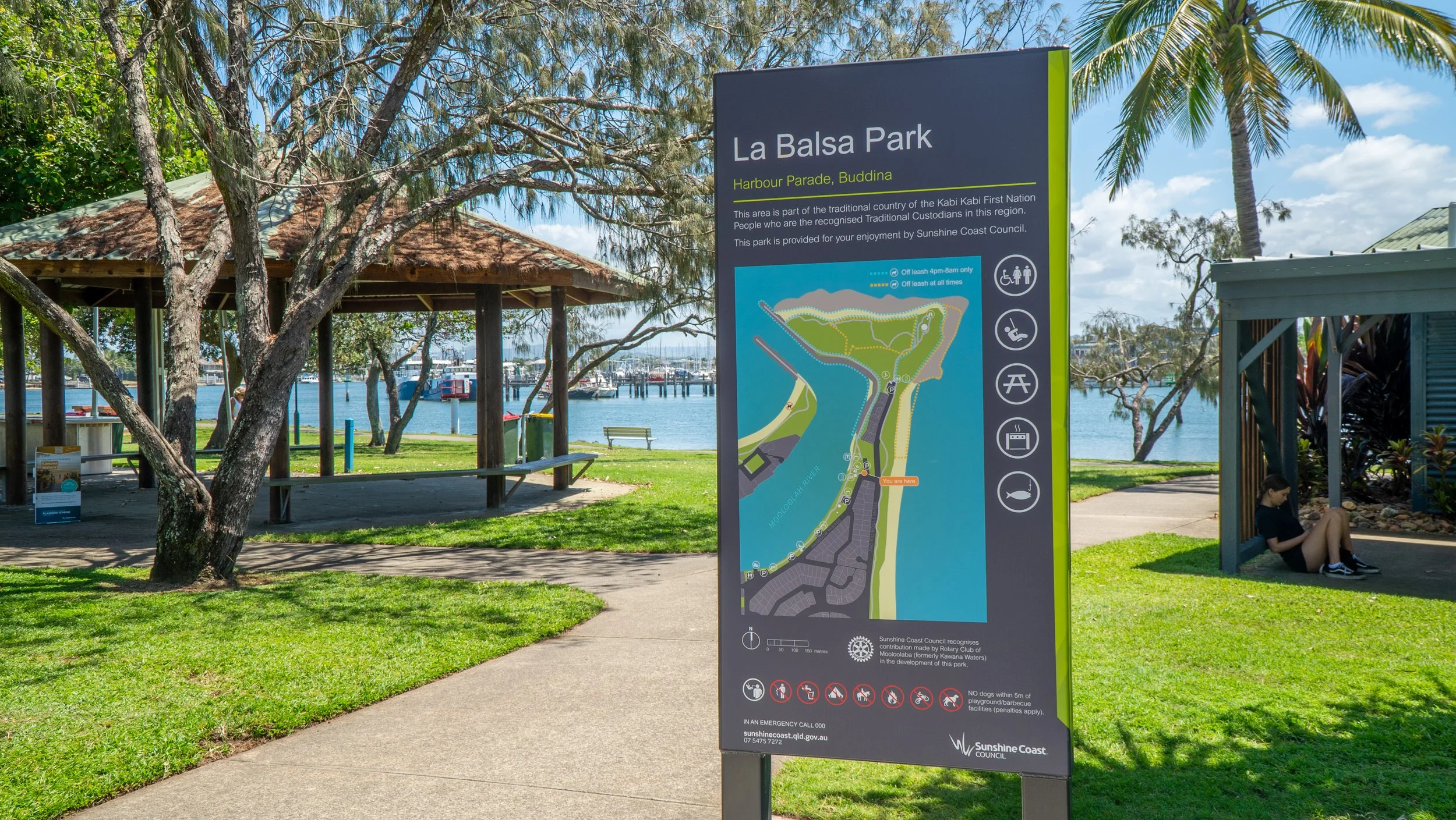 Signboard at La Balsa Park in Harbour Parade, Buddina, with a map of the park and recreational icons, set in a lush green park with trees, a pathway, a gazebo, and the water in the background.