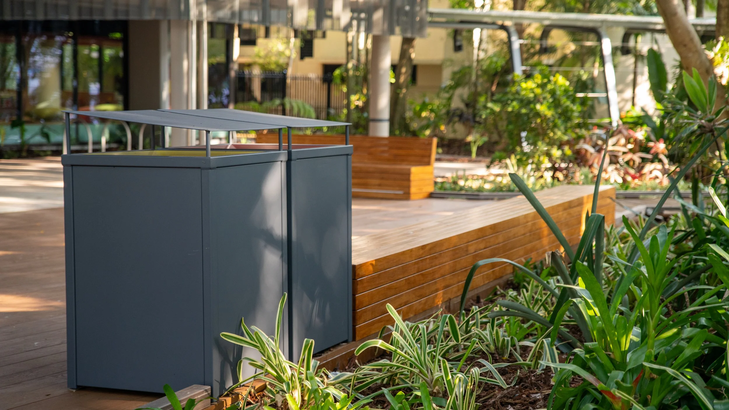 Outdoor urban park area with green plants, a blue trash bin, wooden benches, and a playground in the background.