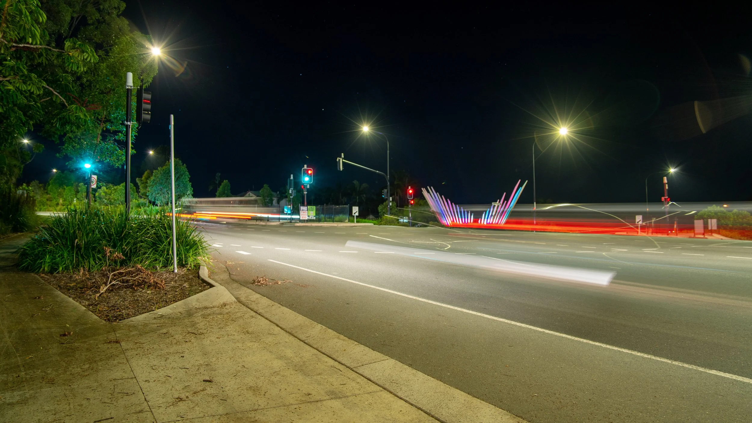 Nighttime city intersection with traffic lights, illuminated street signs, and light trails from passing vehicles, with trees and greenery on the sidewalk.