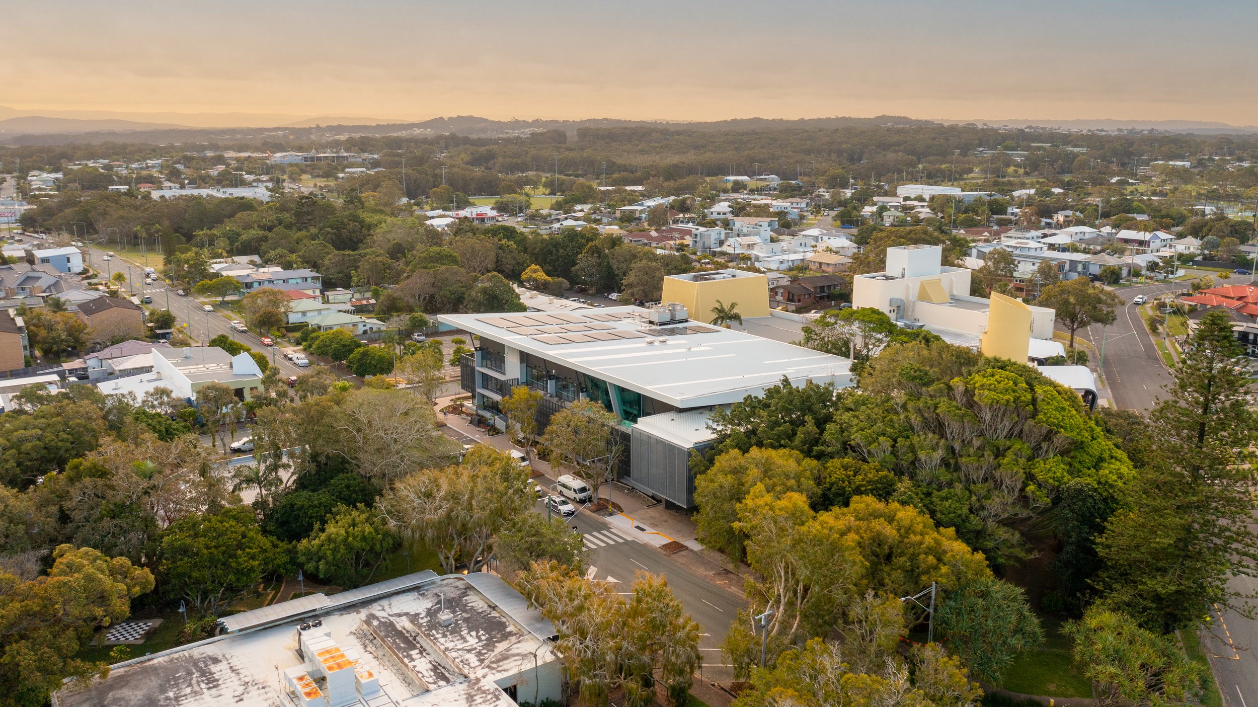 An aerial view of a cityscape during sunset, featuring a modern building surrounded by trees and residential houses.