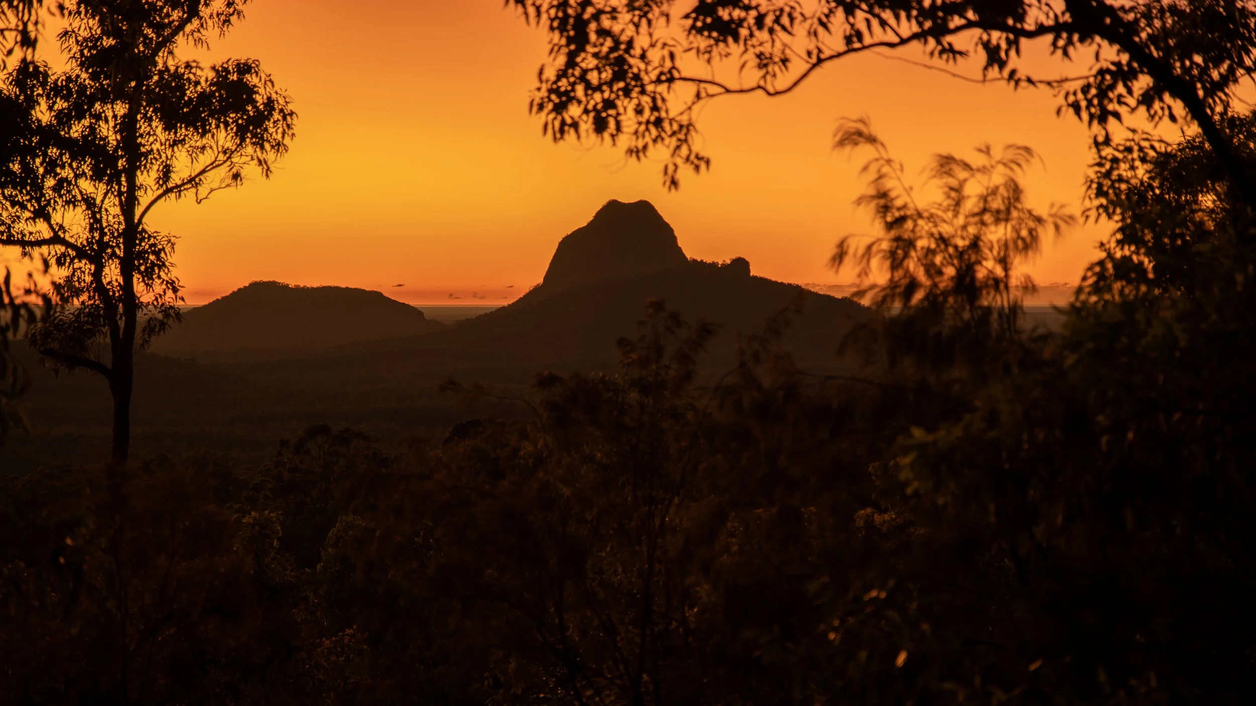 Sunset over a silhouette of mountains and trees with a vibrant orange sky.
