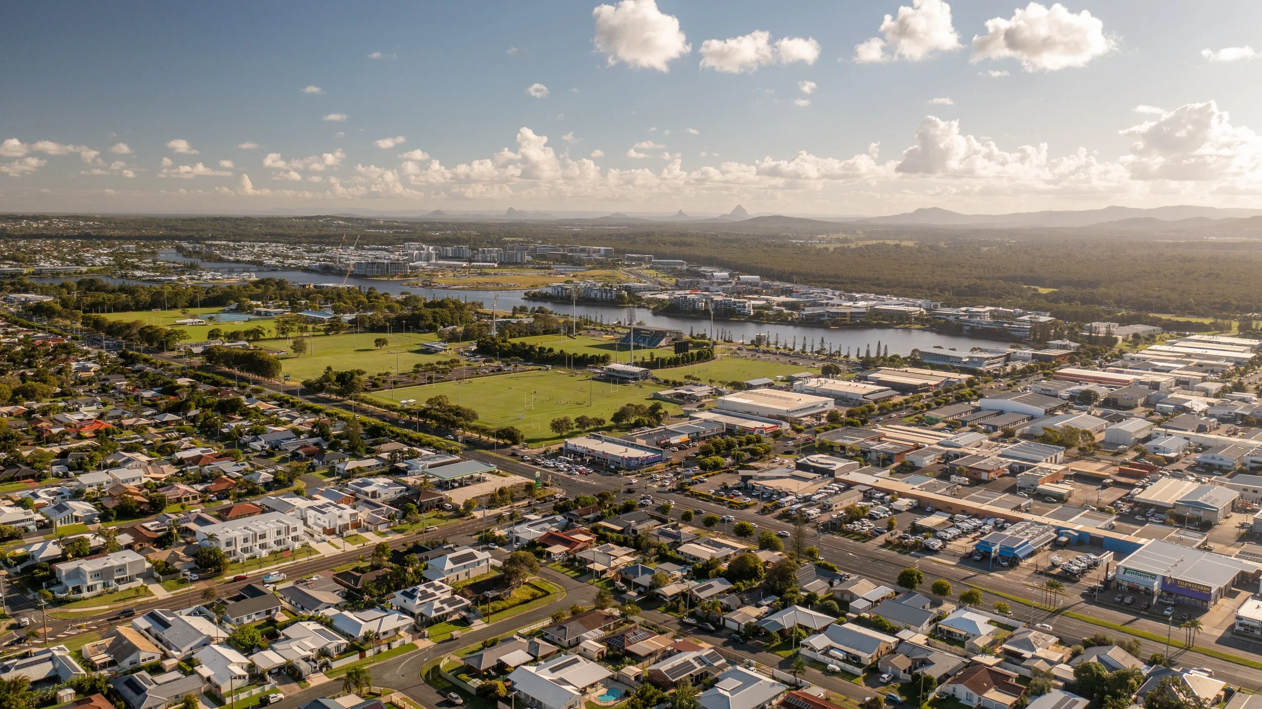 Aerial view of a suburban neighborhood with houses, a park, a river, and commercial buildings under a partly cloudy sky.