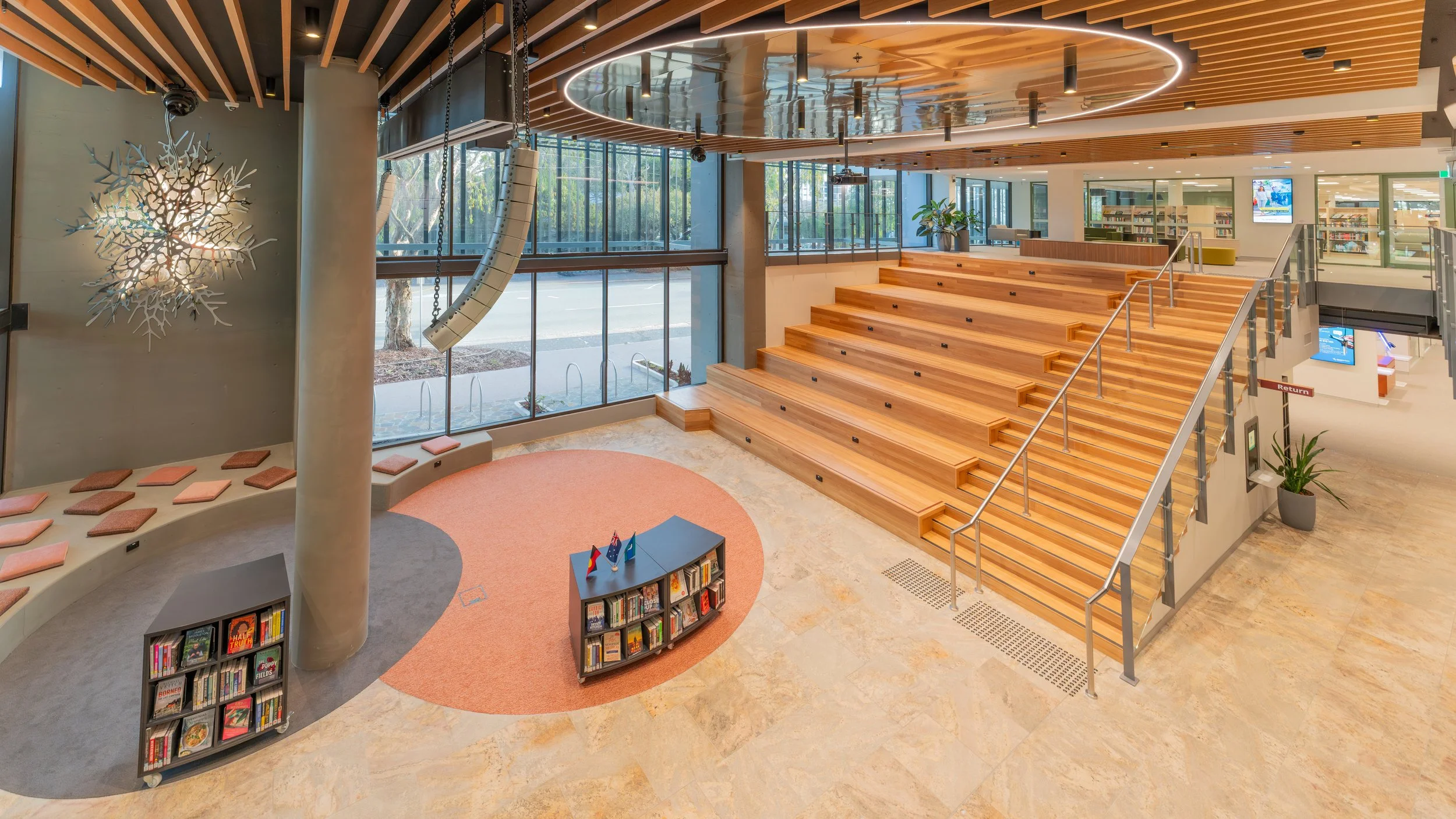 Interior of a modern library with a wooden staircase, large windows, bookshelves, and a seating area with cushions surrounding a pillar. There are flags and books on a small table, and some digital screens.