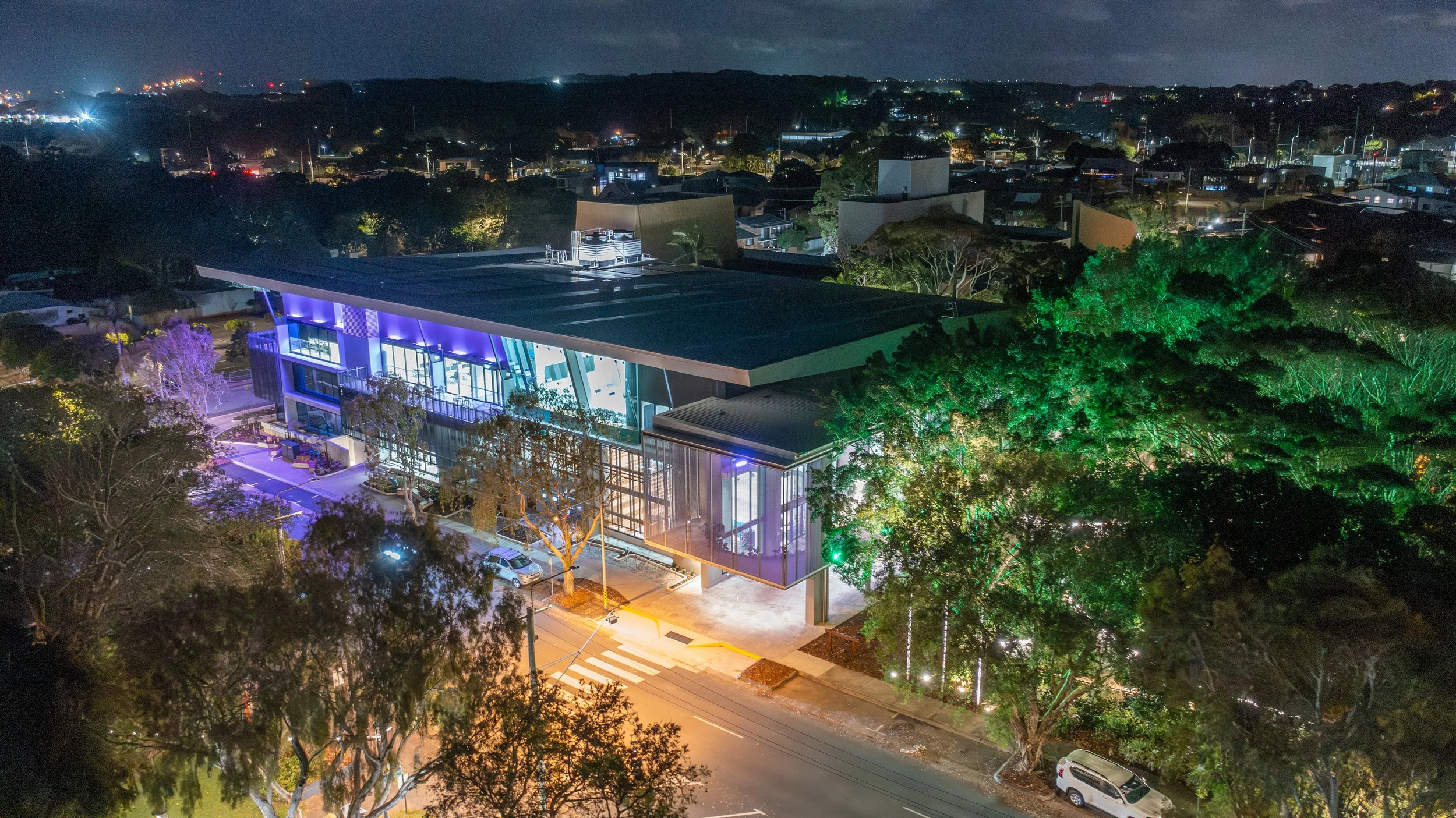 A modern building with colorful lighting is situated among tall trees on a city street at night, with city lights and residential areas in the background.
