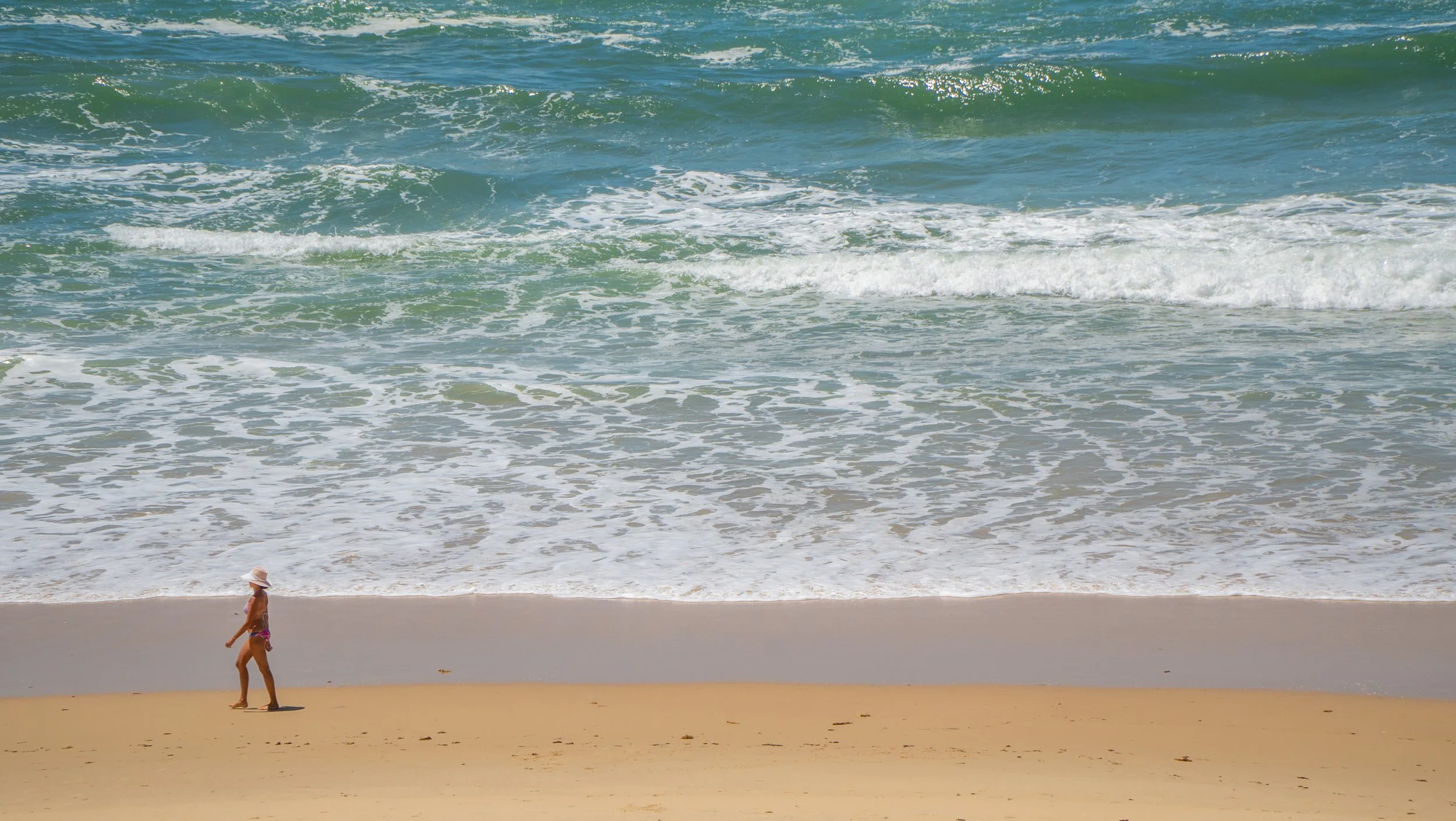 A woman walking on a sandy beach near the ocean waves with turquoise water and white foam.