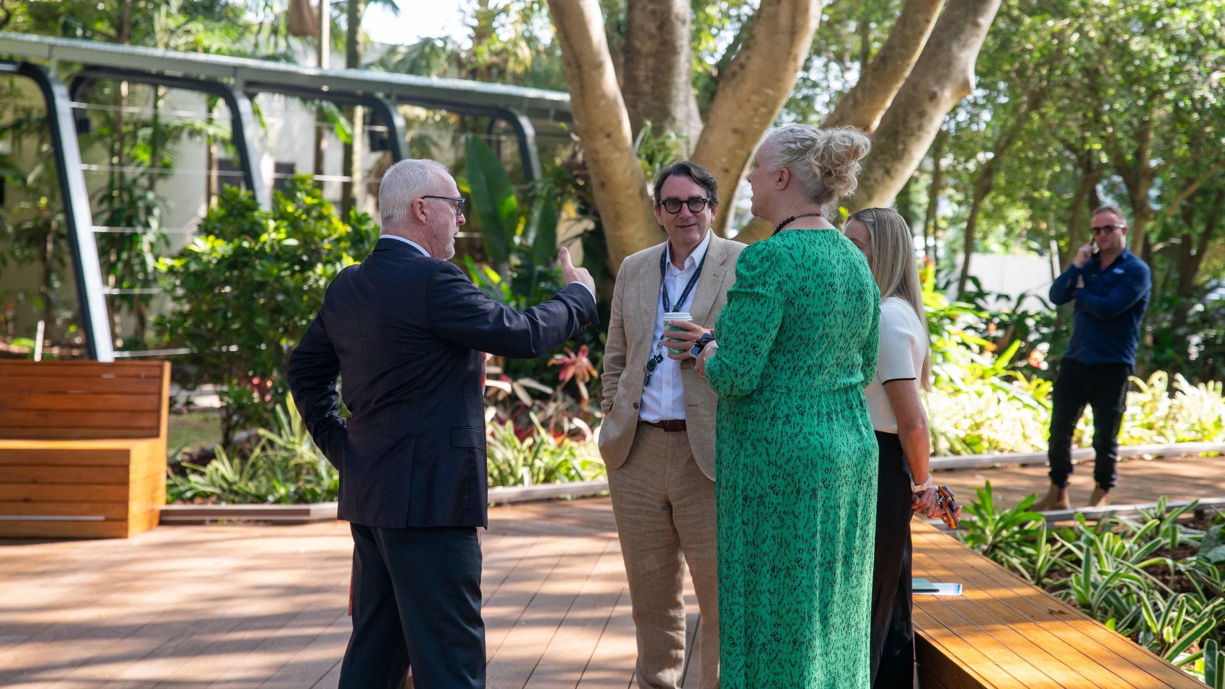 Four people engaged in conversation outdoors in a garden setting, with another person standing in the background on a phone call.