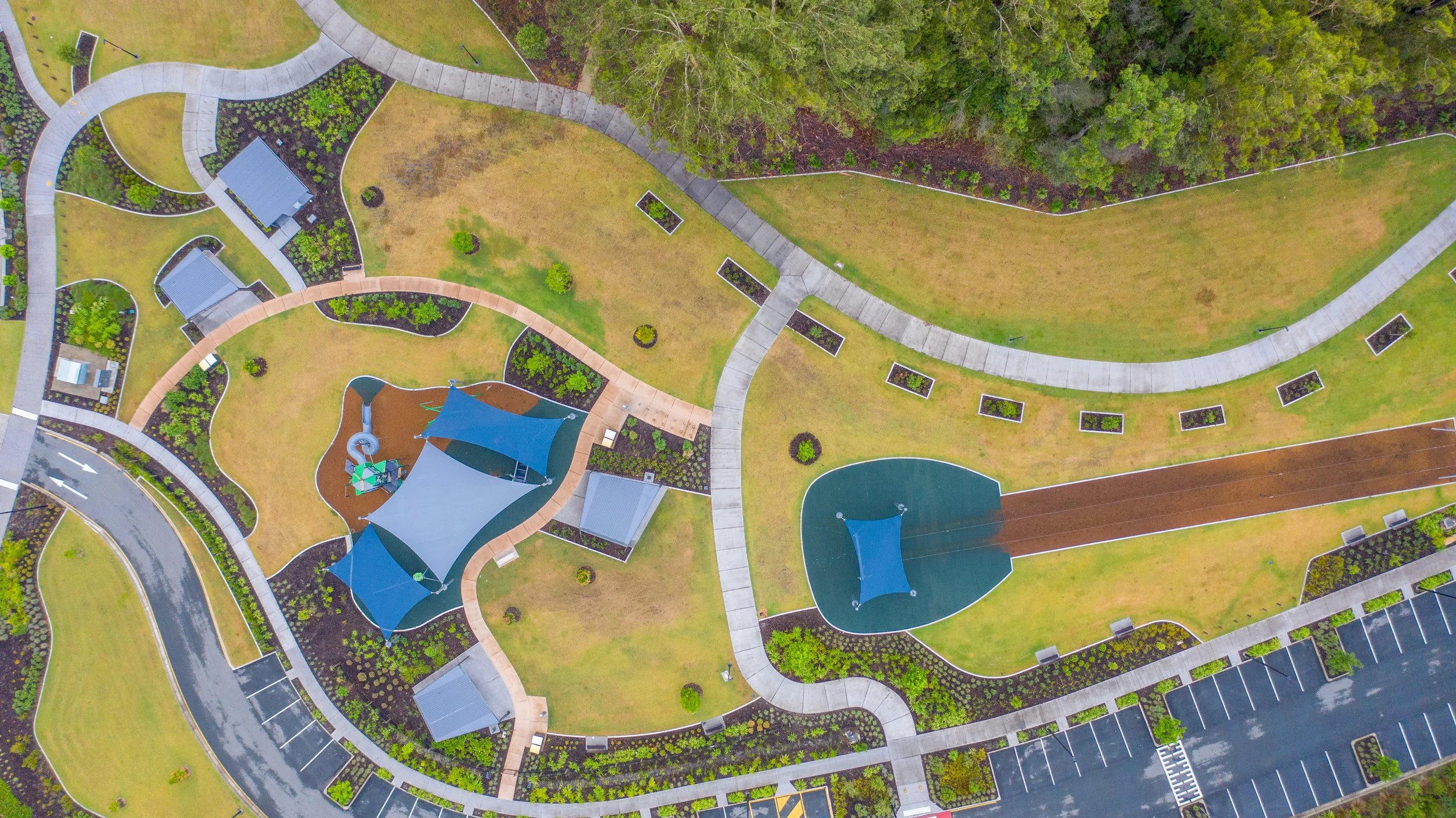 An aerial view of a landscaped public park with walking paths, small covered structures, a playground with shade sails, a basketball court, and surrounding parking lot.