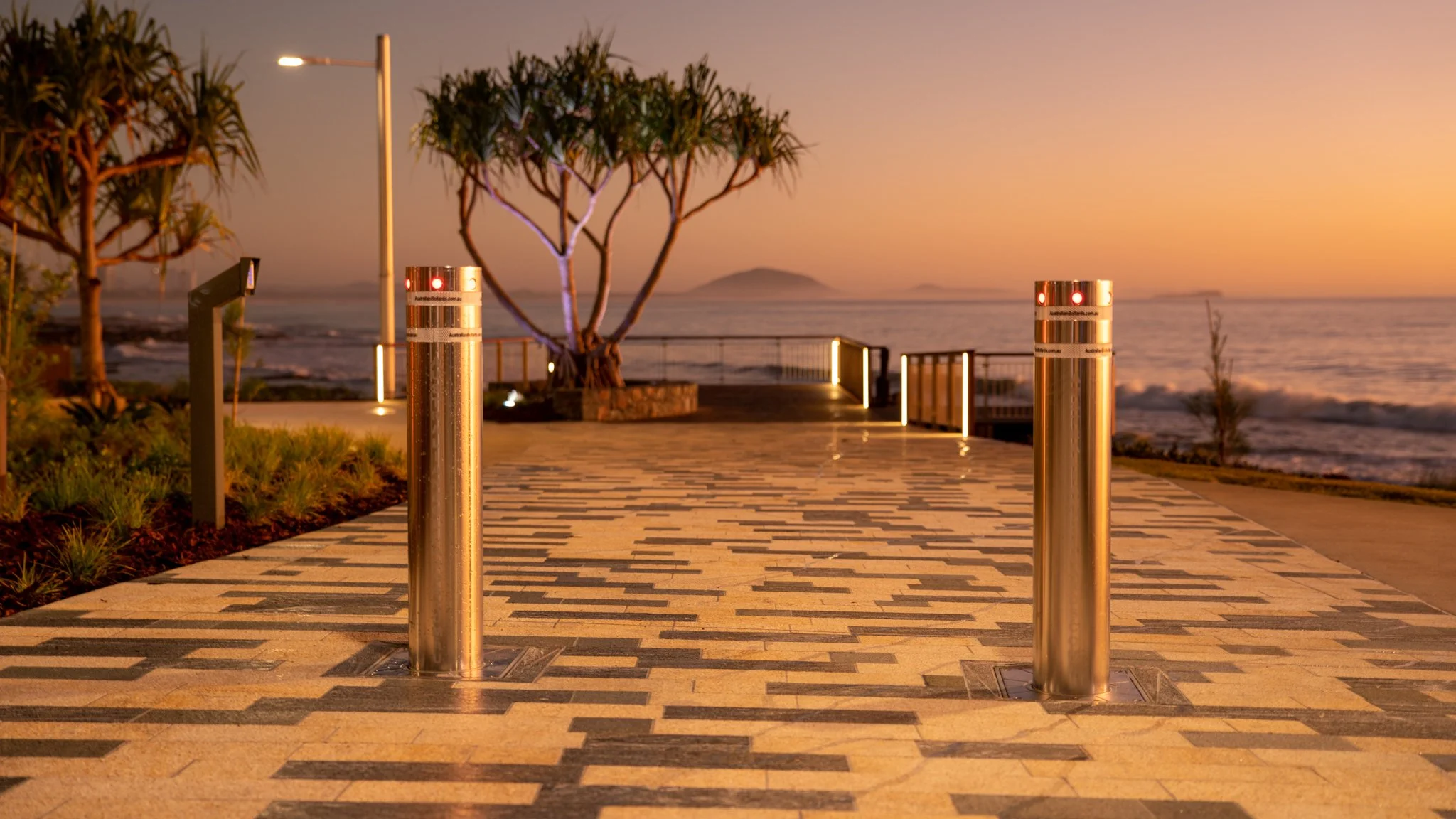 Empty seaside walkway at sunset with stainless steel turnstiles, trees, and ocean in background.