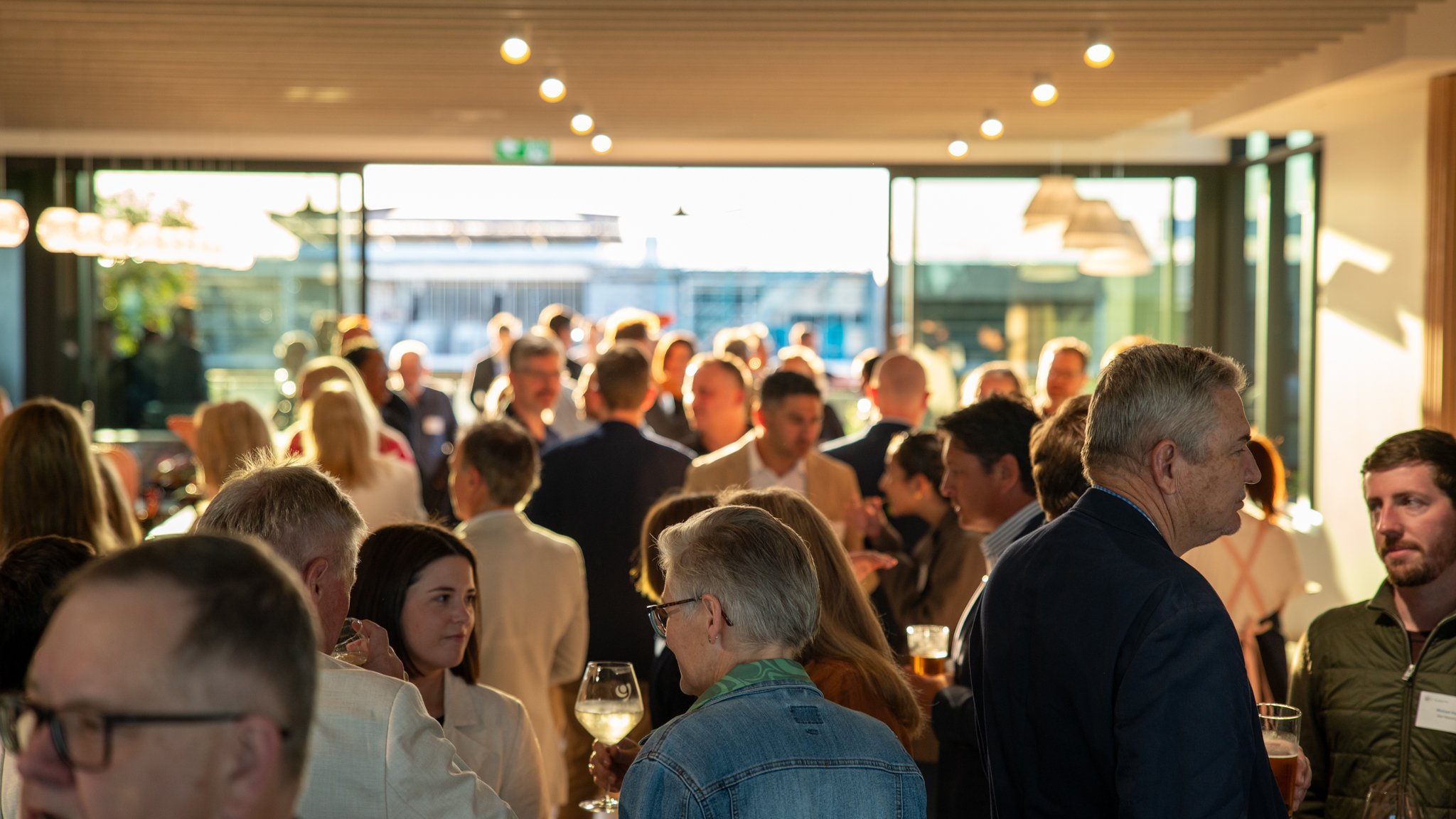 People attending a social gathering or party indoors with sunlight streaming in, some holding drinks, engaging in conversations, and mingling.