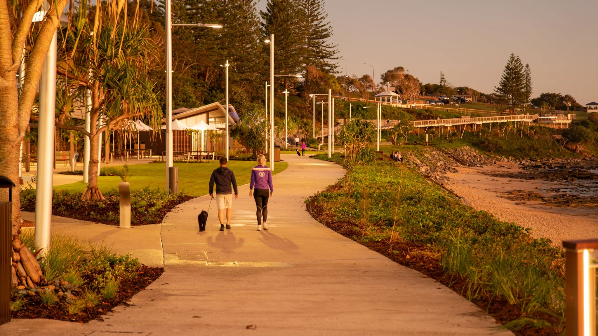 People walking along a scenic coastal path during sunset, with palm trees and a rocky beach on the right, and a lush green lawn with trees on the left.