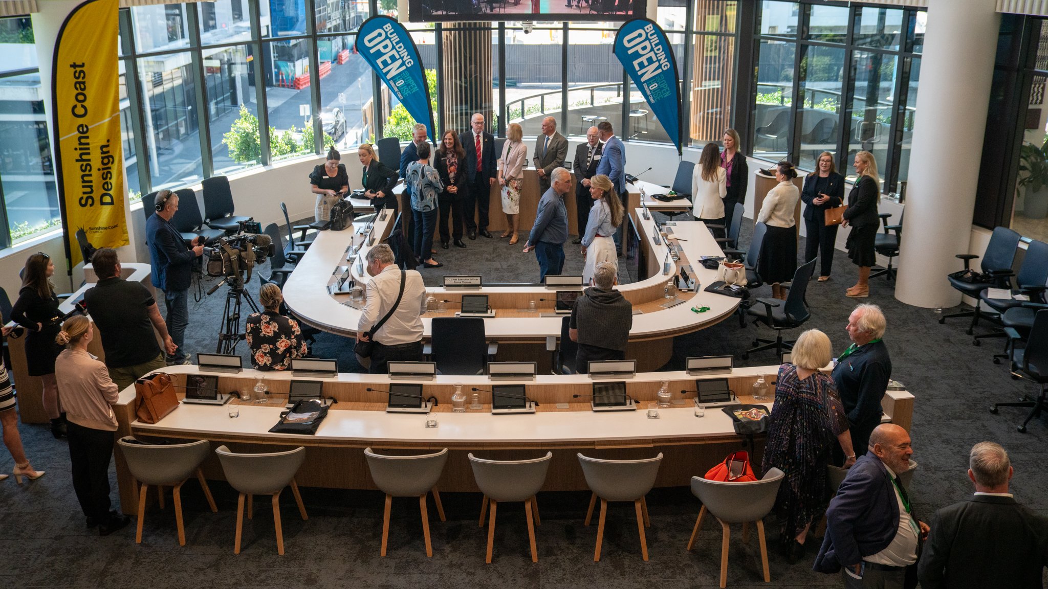 A meeting room filled with numerous people, some standing and chatting, others seated at a curved conference table with laptops. There are banners on flags that say 'Building Open' and a yellow banner on the left that reads 'Sunshine Coast Design.' T