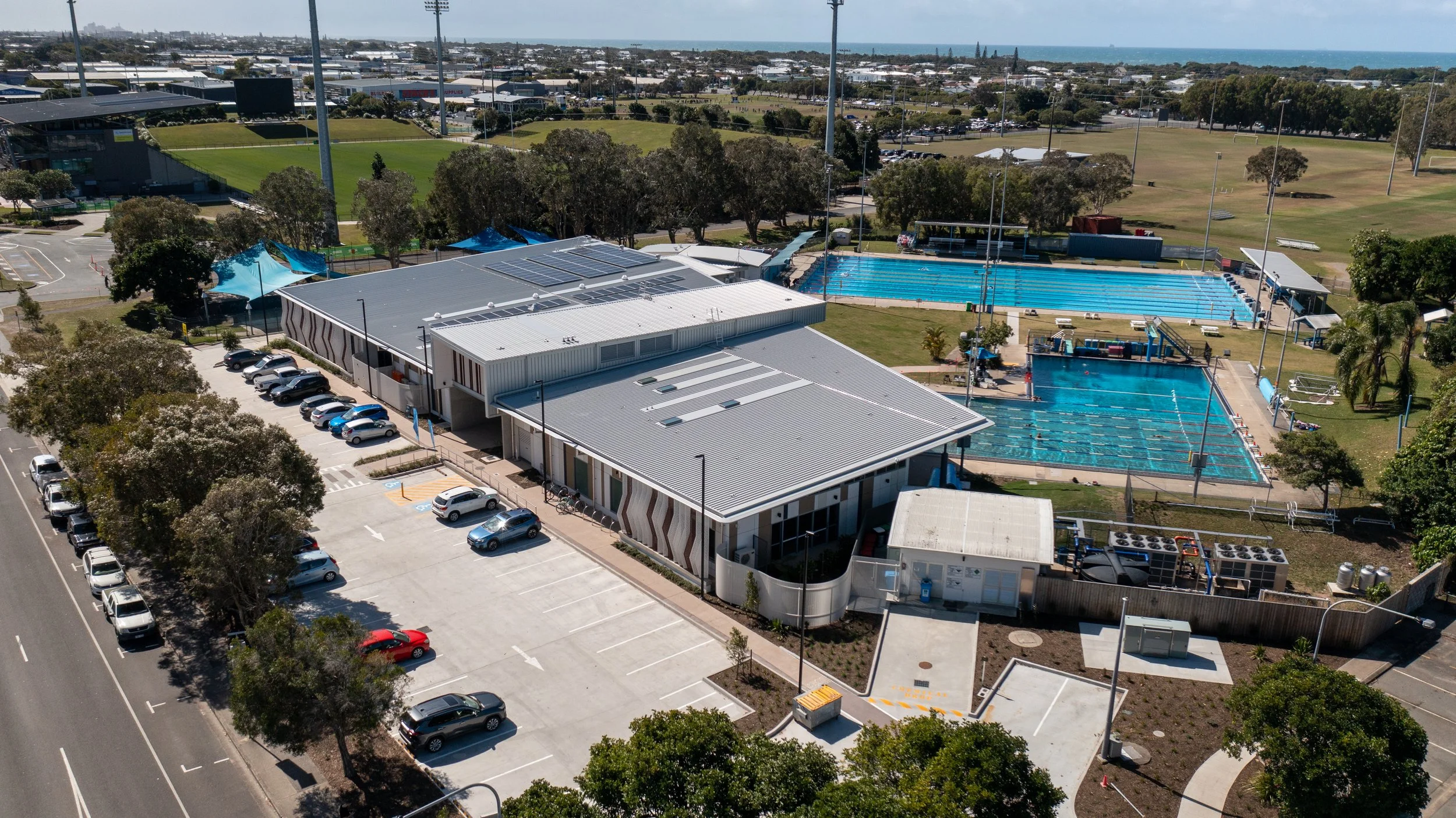An aerial view of a sports complex with swimming pools, parking lot, and a grassy field, possibly for a recreational or aquatic center.