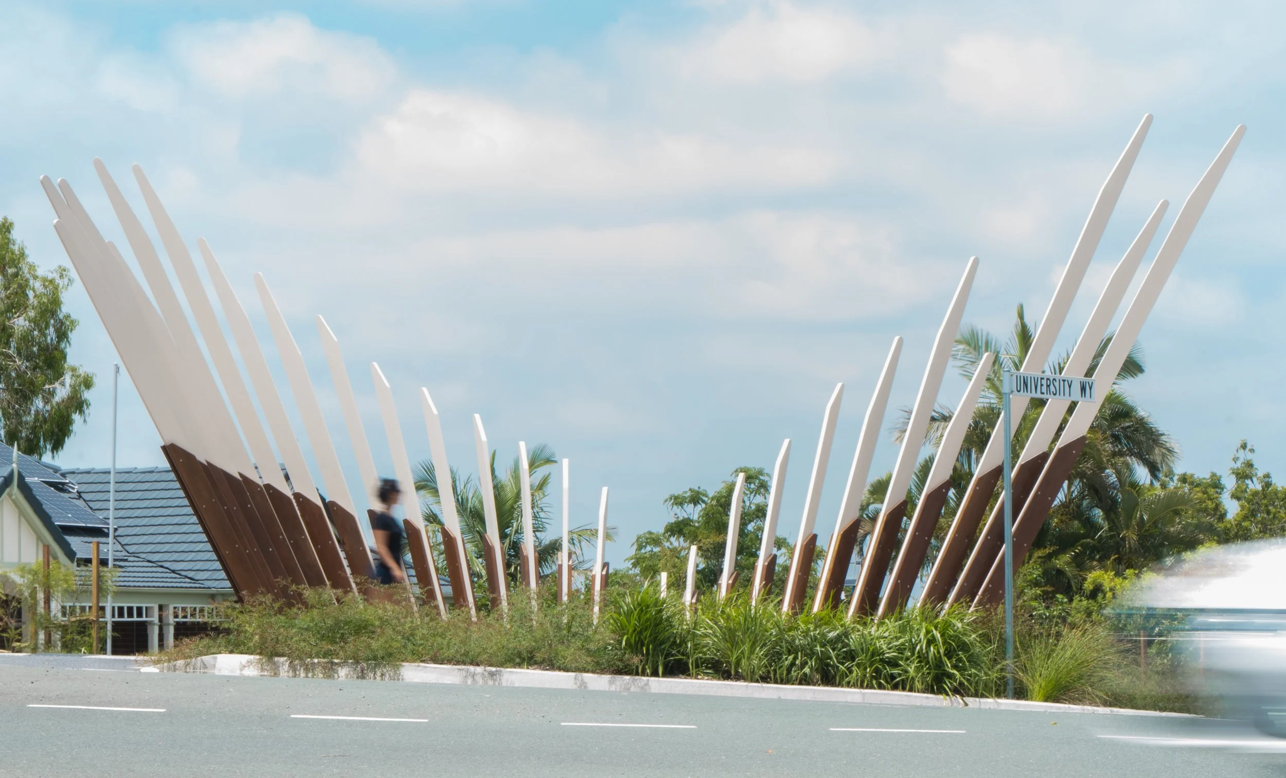 Modern architectural sculpture with white and brown vertical slats, landscaping, and a street sign reading 'University Wy' in a sunny suburban area.