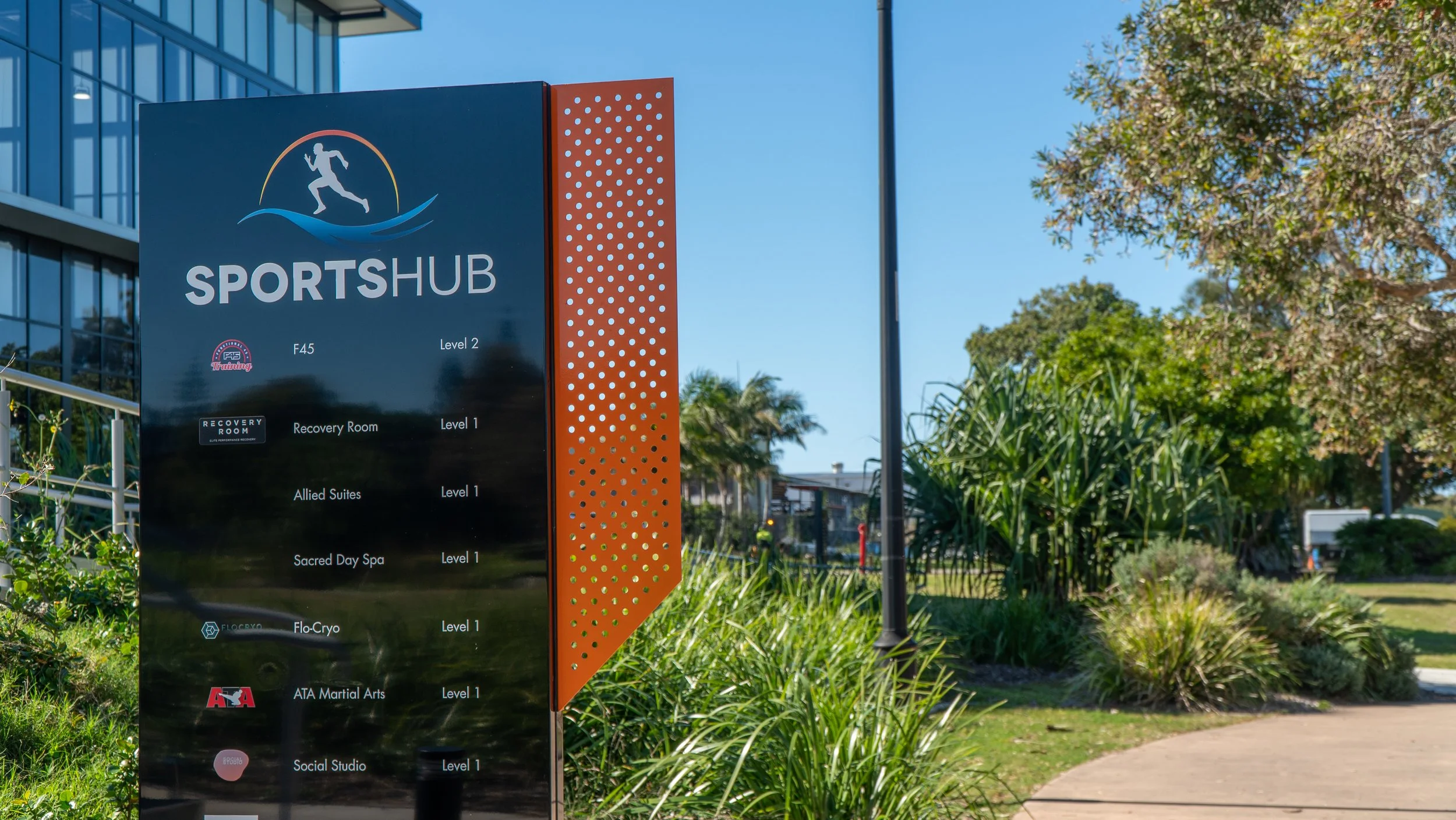 Outdoor directory sign for Sportshub, listing facilities such as F45 Training, Recovery Room, Allied Suites, Sacred Day Spa, Flo-Cryo, ATA Martial Arts, and Social Studio with floor levels indicated, set against a landscaped background with trees and