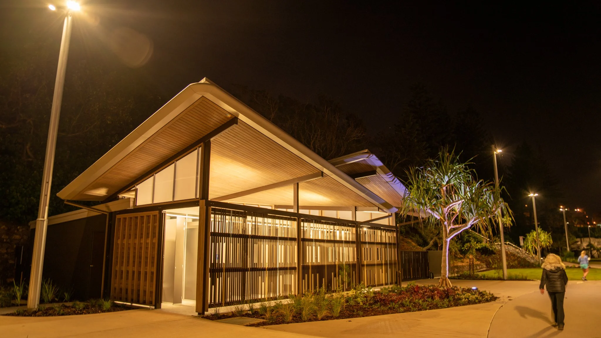 Night view of a modern building with a sloped roof, illuminated from inside, with a wooden and metal fence, trees, and a pathway with people walking.
