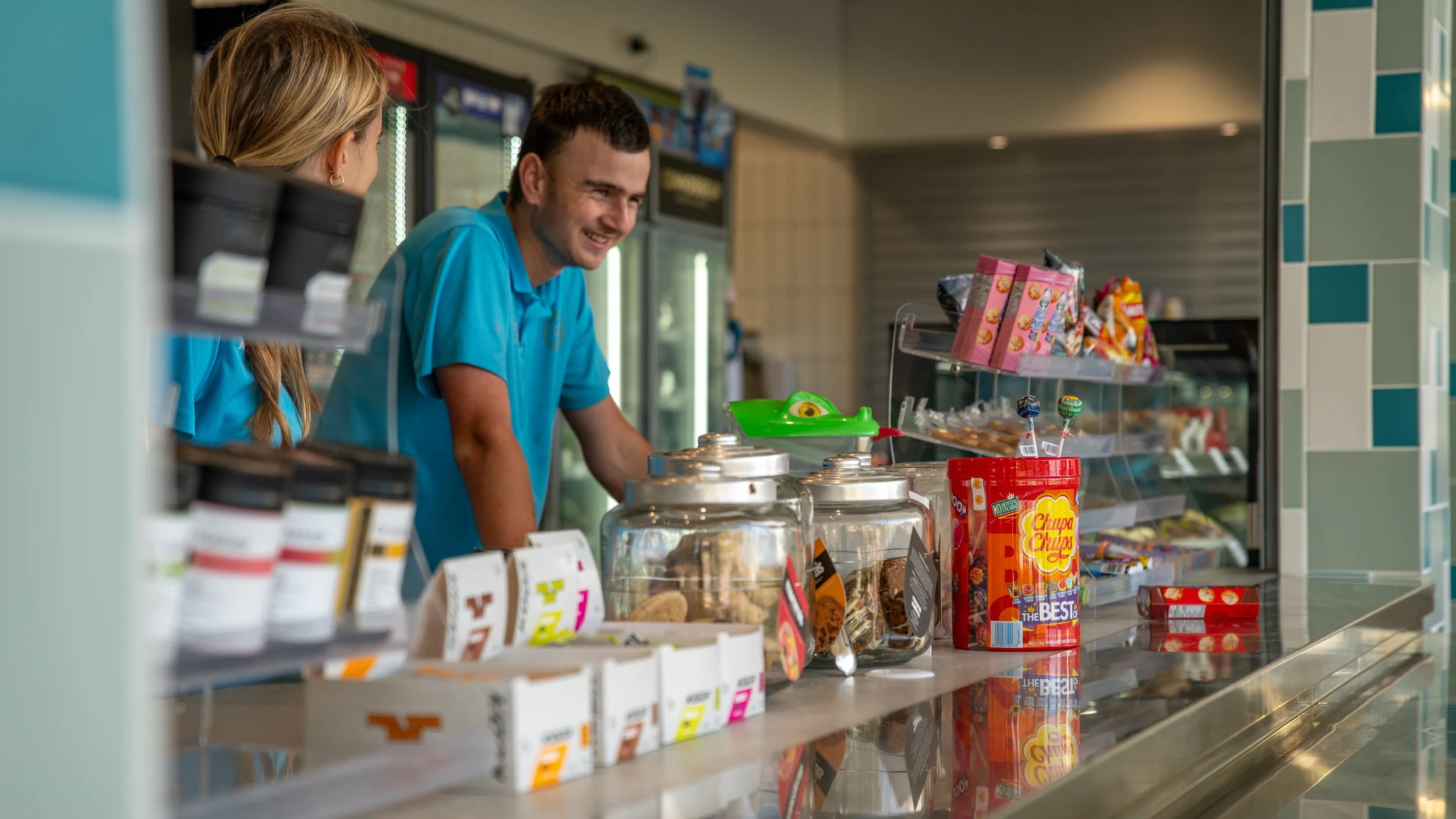 A man and woman working behind a snack counter in a convenience store, with various candies, snacks, and jars of cookies on the counter.