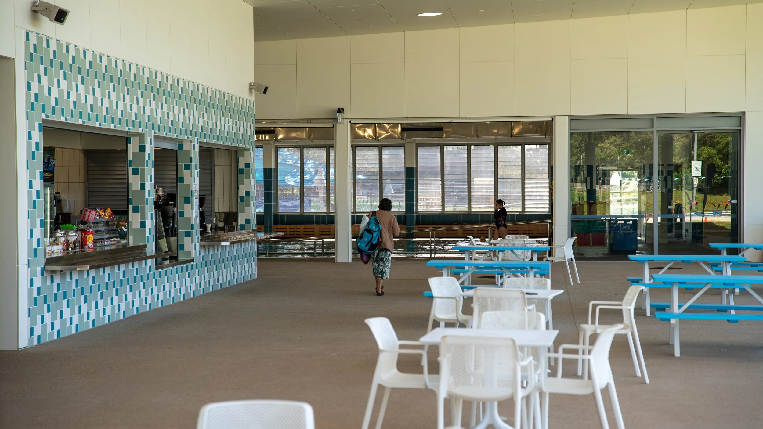 Indoor swimming pool area with a snack bar, seating, and a few people, including a woman walking towards the pool, and a person near the pool.