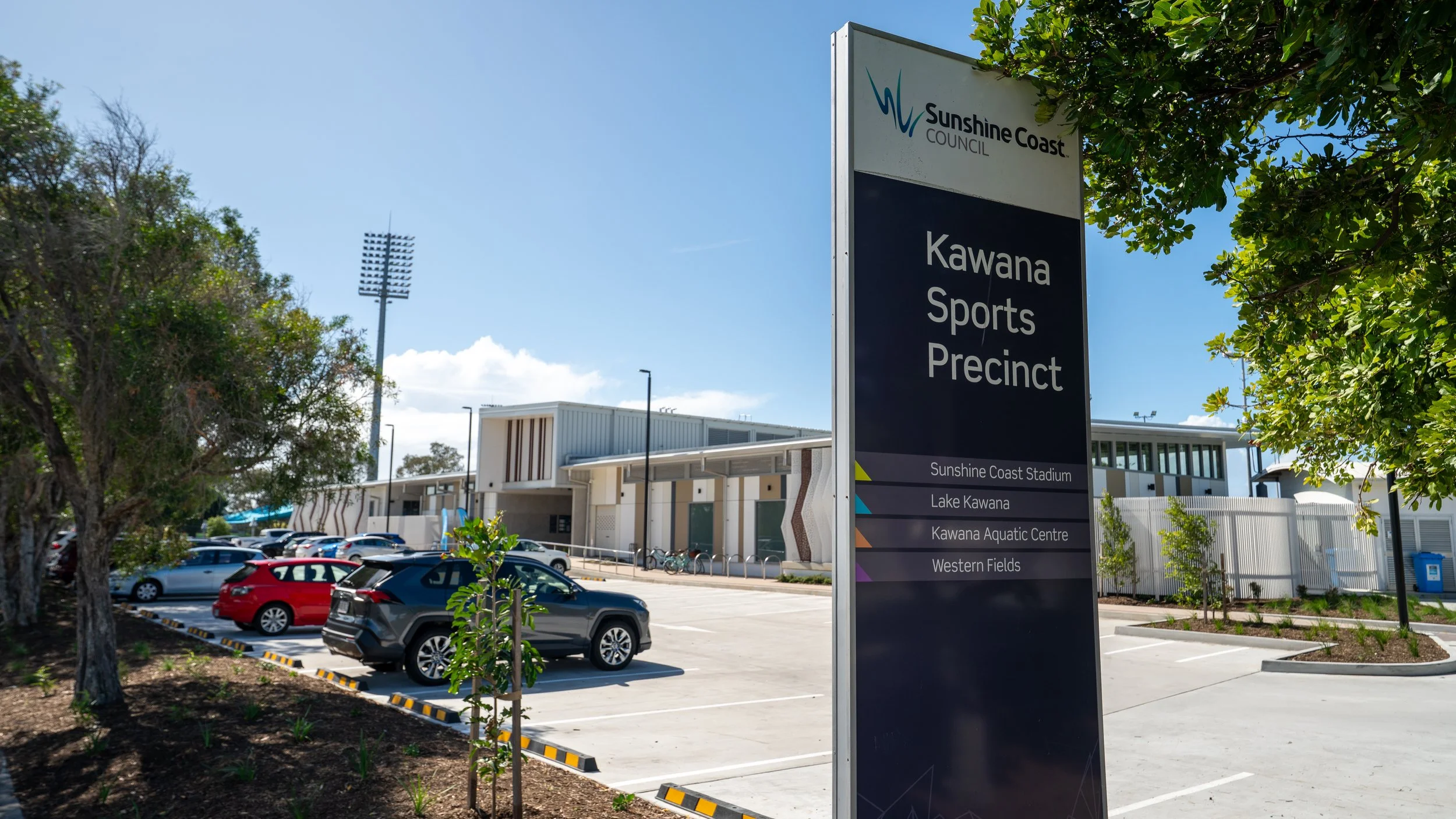 Parking lot outside the Kawana Sports Precinct with a large sign providing directions to Sunshine Coast Stadium, Lake Kawana, Kawana Aquatic Centre, and Western Fields.