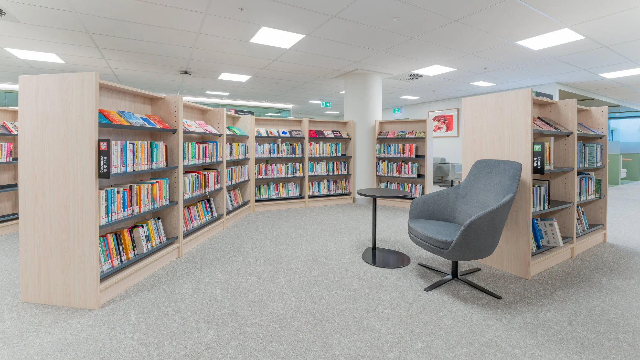 A quiet corner inside a modern library with wooden bookshelves filled with colorful books, a gray lounge chair, and a small round black table on a light gray carpet.