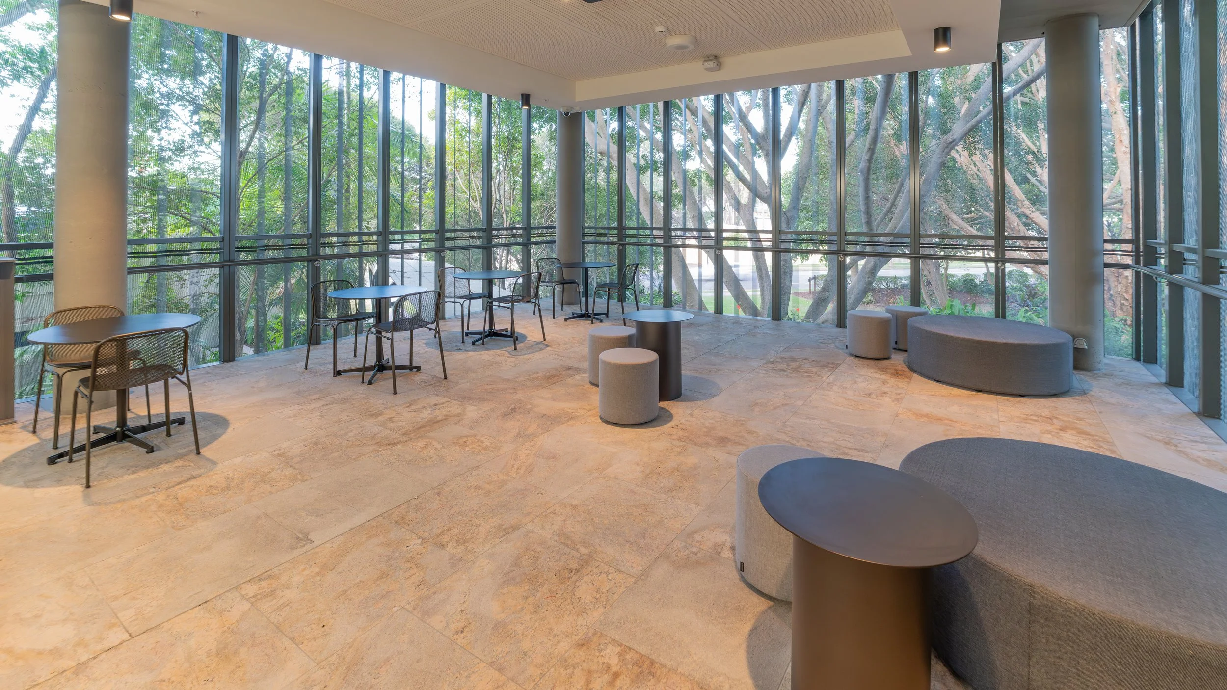 An empty modern indoor space with large floor-to-ceiling windows revealing trees outside. The room has round tables with chairs and cushioned seating, creating a relaxing atmosphere.