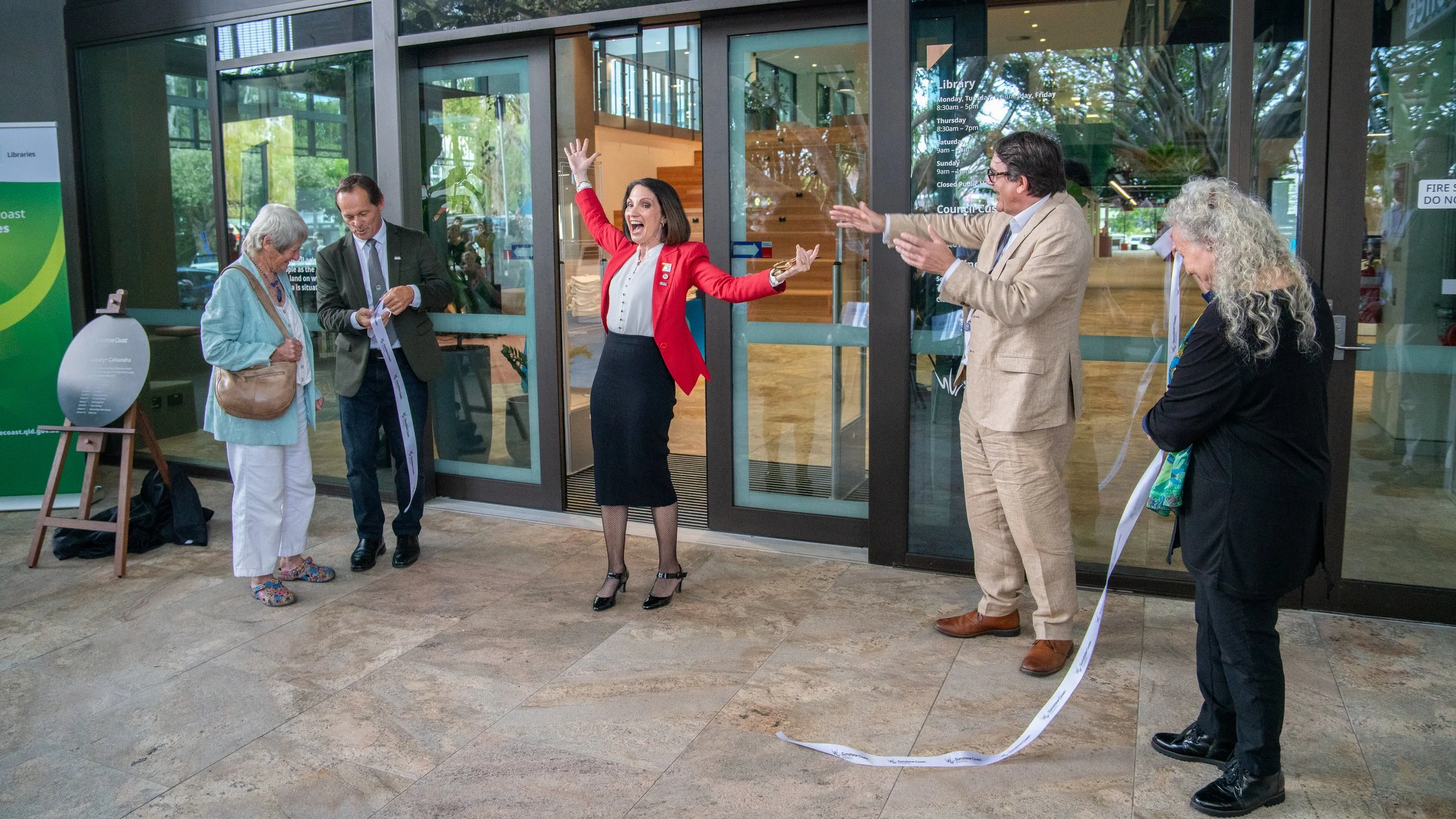 Group of five people at the entrance of a building celebrating a ribbon-cutting event, with one woman in a red blazer appearing excited and others clapping or holding the ribbon.