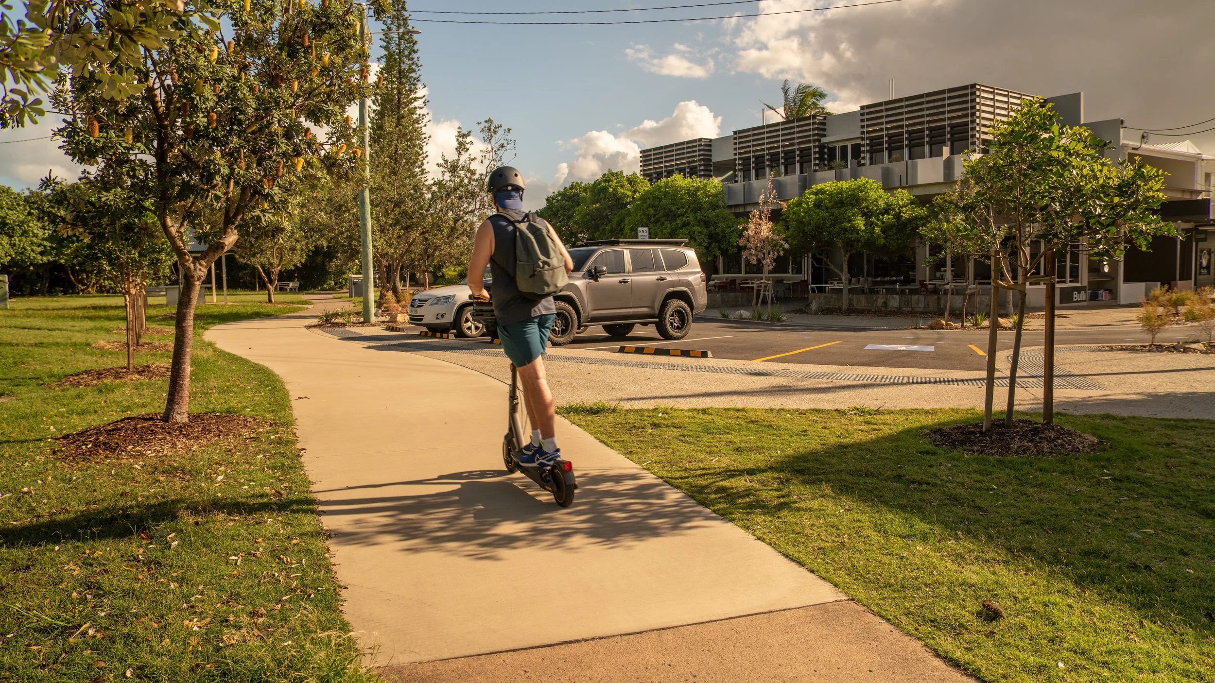 A person riding an electric scooter on a sidewalk in a park with trees, grass, and parked cars in the background during late afternoon sunlight.