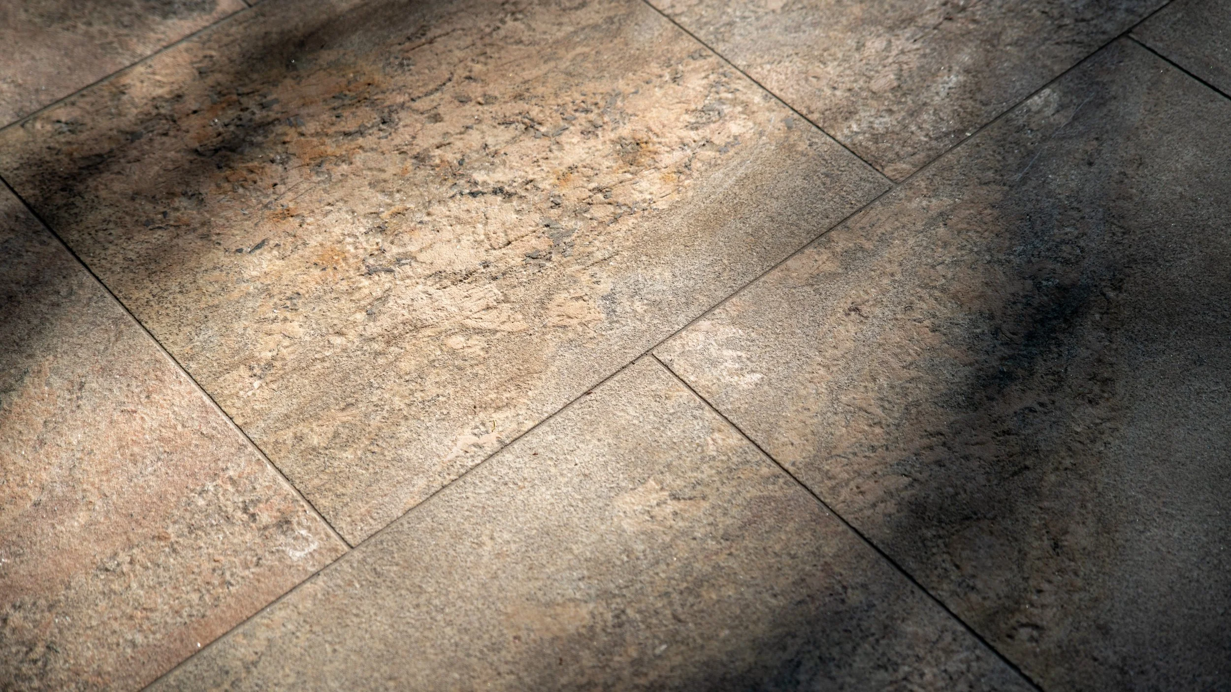 Close-up of a tiled stone floor with shades of brown, tan, and gray, showing grout lines and texture.