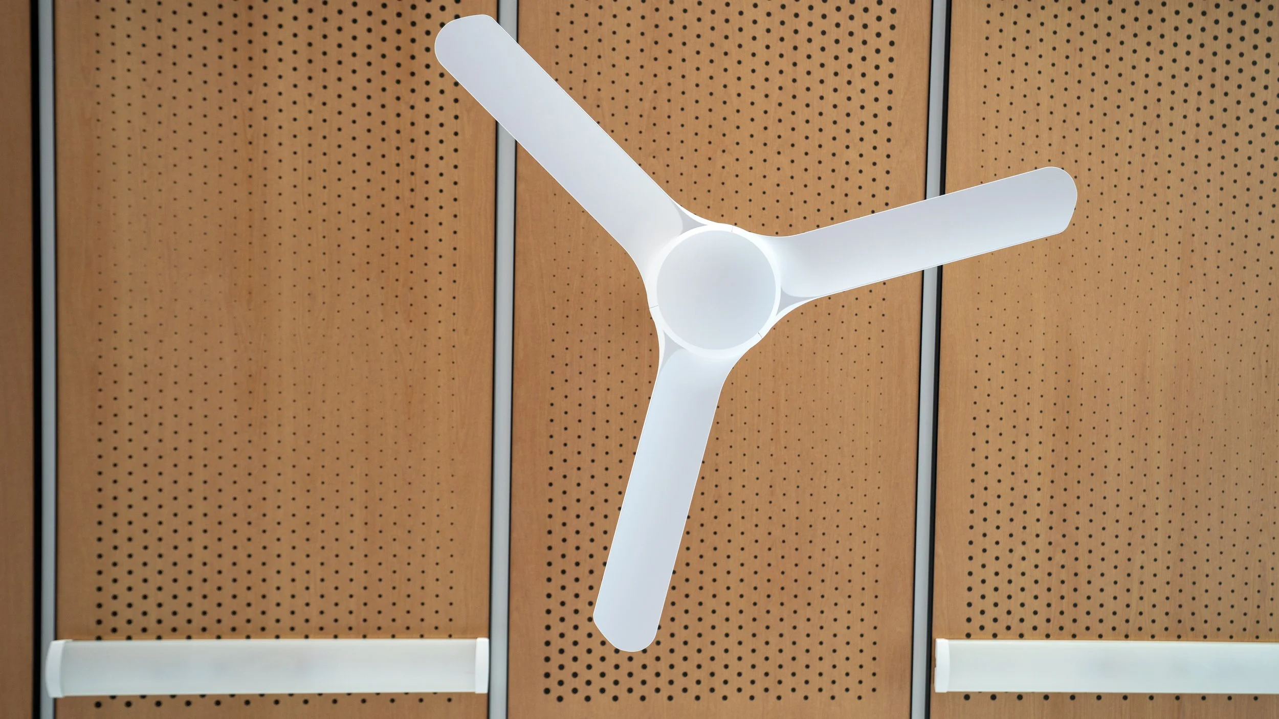 White ceiling fan with three blades mounted on a perforated wooden ceiling with vertical metal beams.